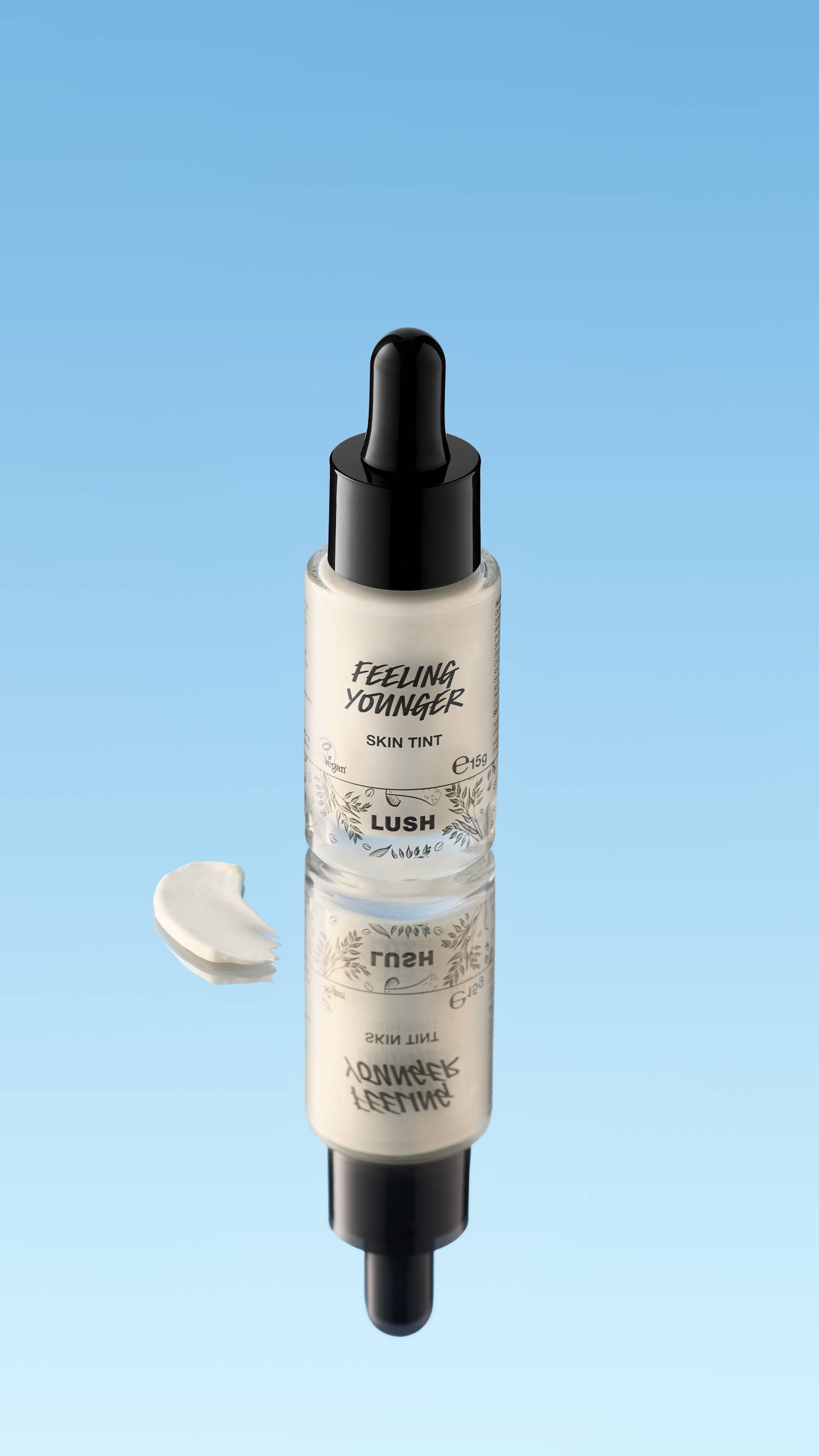 An angled product shot of a dropper bottle of LUSH Feeling Younger Skin Tint with a black cap, standing on a reflective surface. A small, creamy, off-white swatch of the product rests next to the bottle, all against a bright blue background.