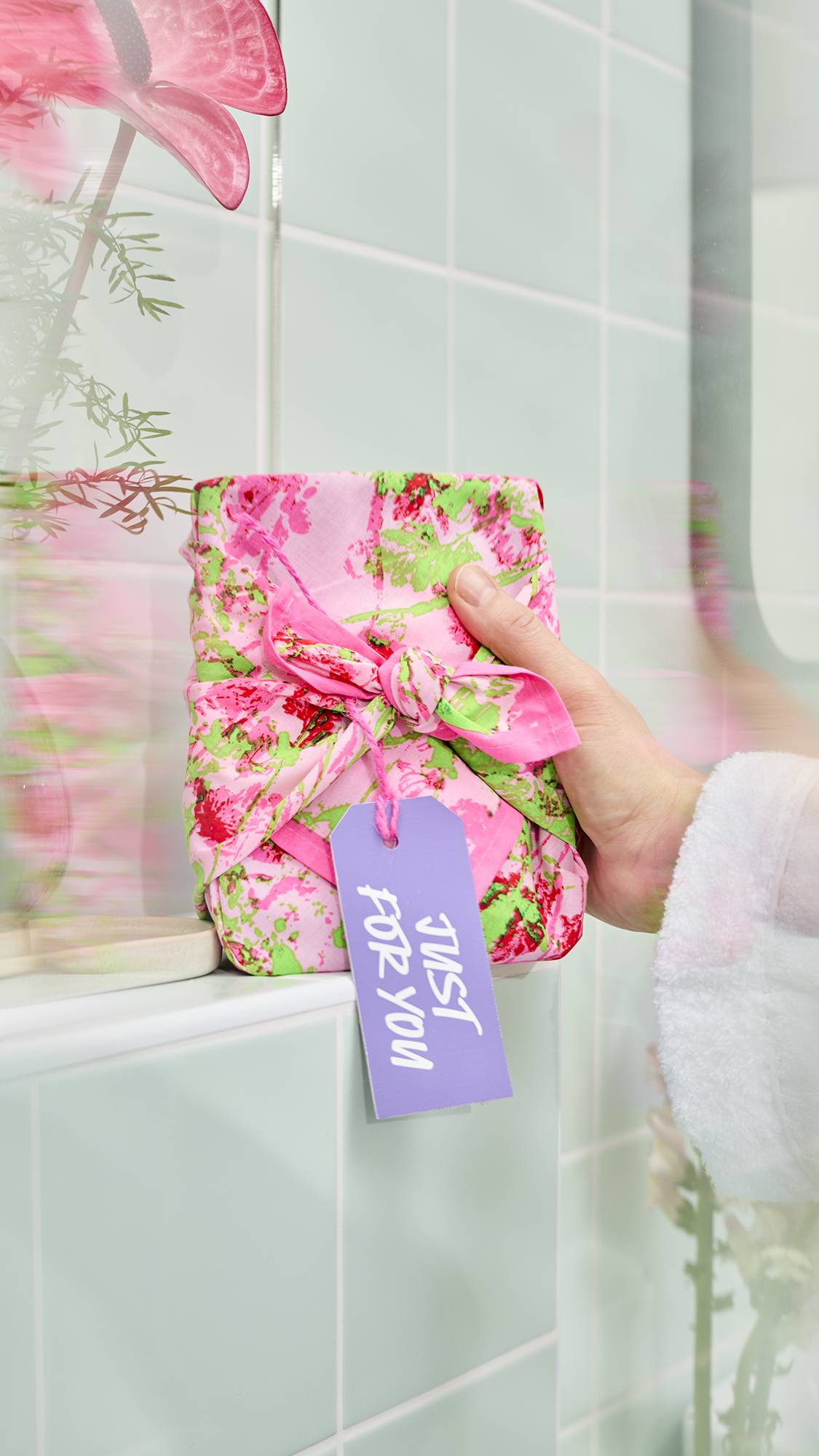 The image shows a model's hand resting on a gift wrapped in a pink "Floral Meadow" fabric knot-wrap. The wrap features an impressionist design of red flowers and green stems on a light pink background, finished with a lilac "Just For You" gift tag.