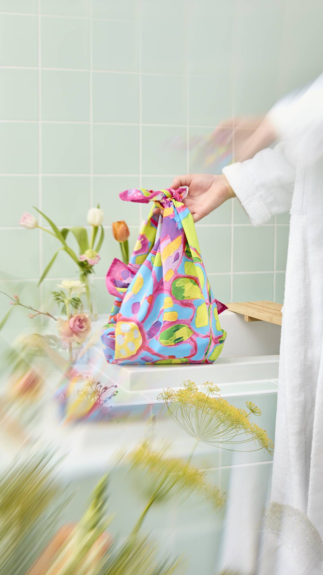 A model's hand holds the colourful "Flourish In Colour" knot-wrap, tied at the top to form a gift bag. The material features a hand-painted floral design in pink, blue, and yellow on a green background, set in a bright, botanical-themed bathroom.