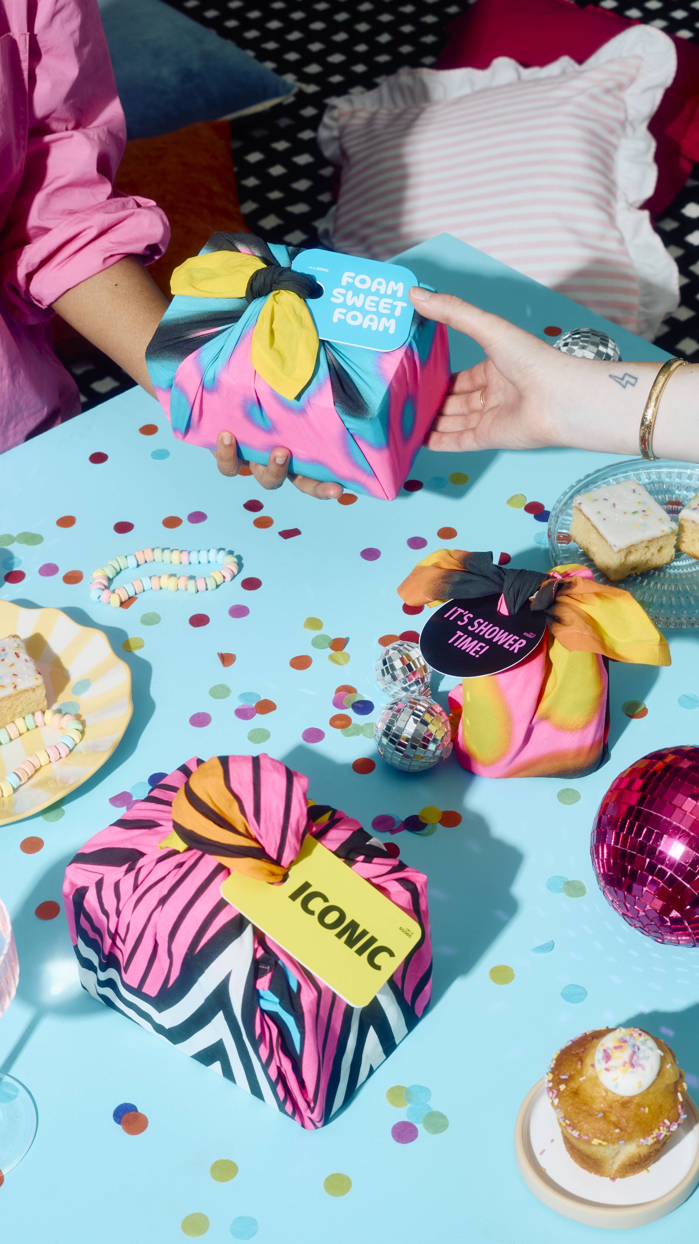 Two hands exchange a fabric-wrapped gift tagged "FOAM SWEET FOAM" across a light blue party table scattered with confetti, cakes, disco balls, and other wrapped gifts tagged "IT'S SHOWER TIME!" and "ICONIC".