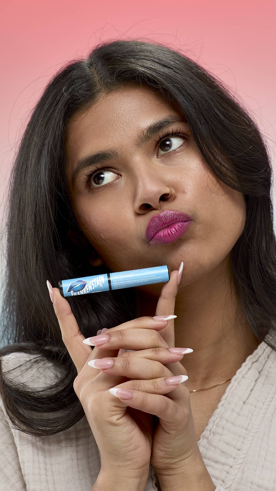 A person with long dark hair holds a light blue tube of Frankenstain lip stain horizontally under their chin. They are wearing the shimmering, deep berry-purple stain on their lips and looking upwards with a playful pout.