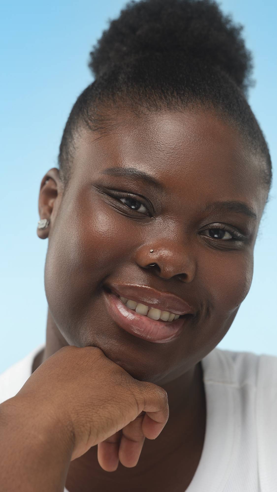 A close-up portrait of a smiling person with deep skin, dark hair in a bun, and a nose stud. Their chin rests on their hand, and their luminous skin, prepped with the set primer, is against a bright blue background.