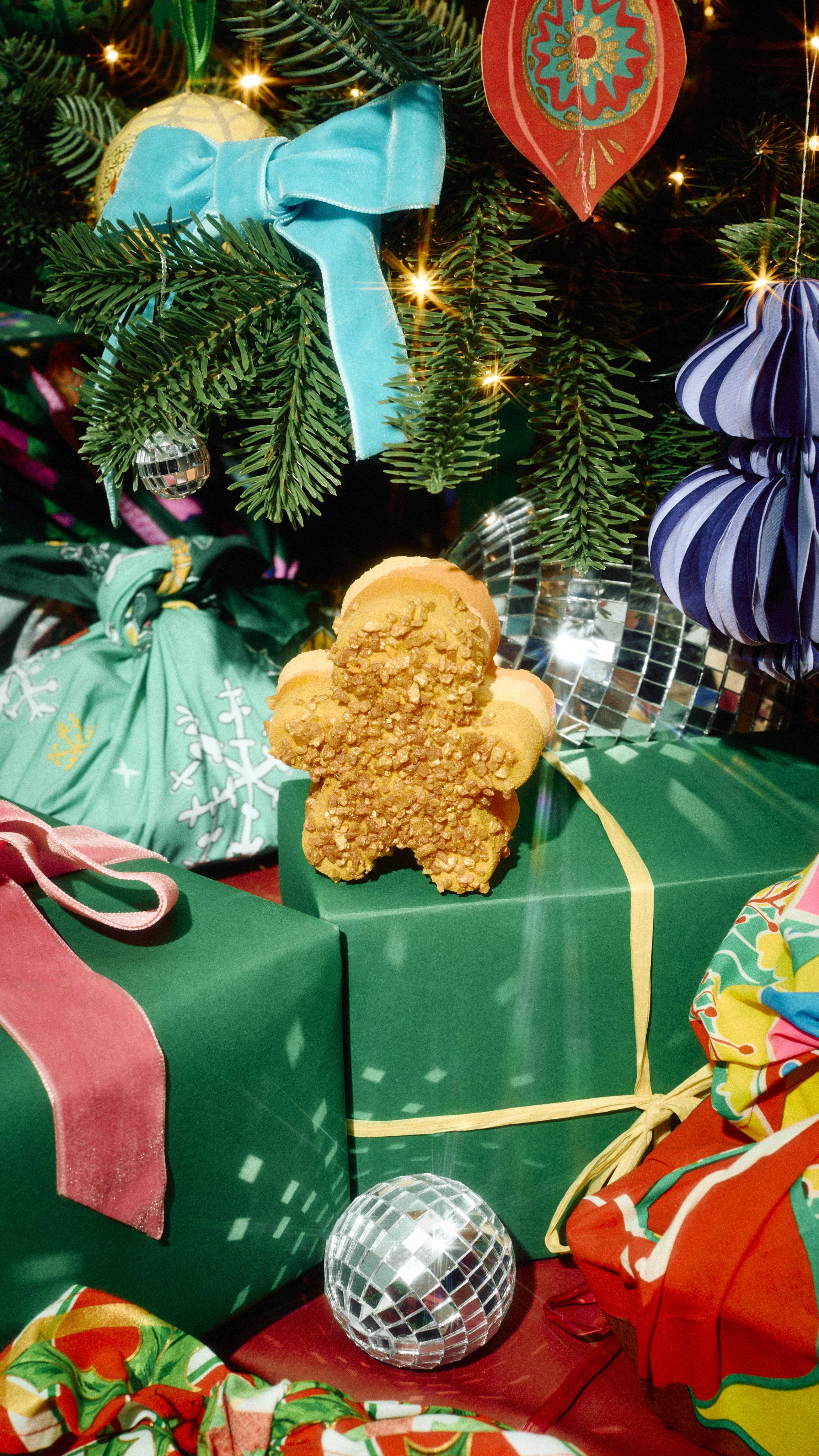 A gingerbread-shaped bath bomb coated in coarse golden crystals rests on a green wrapped present under a decorated Christmas tree with disco balls and velvet ribbons.