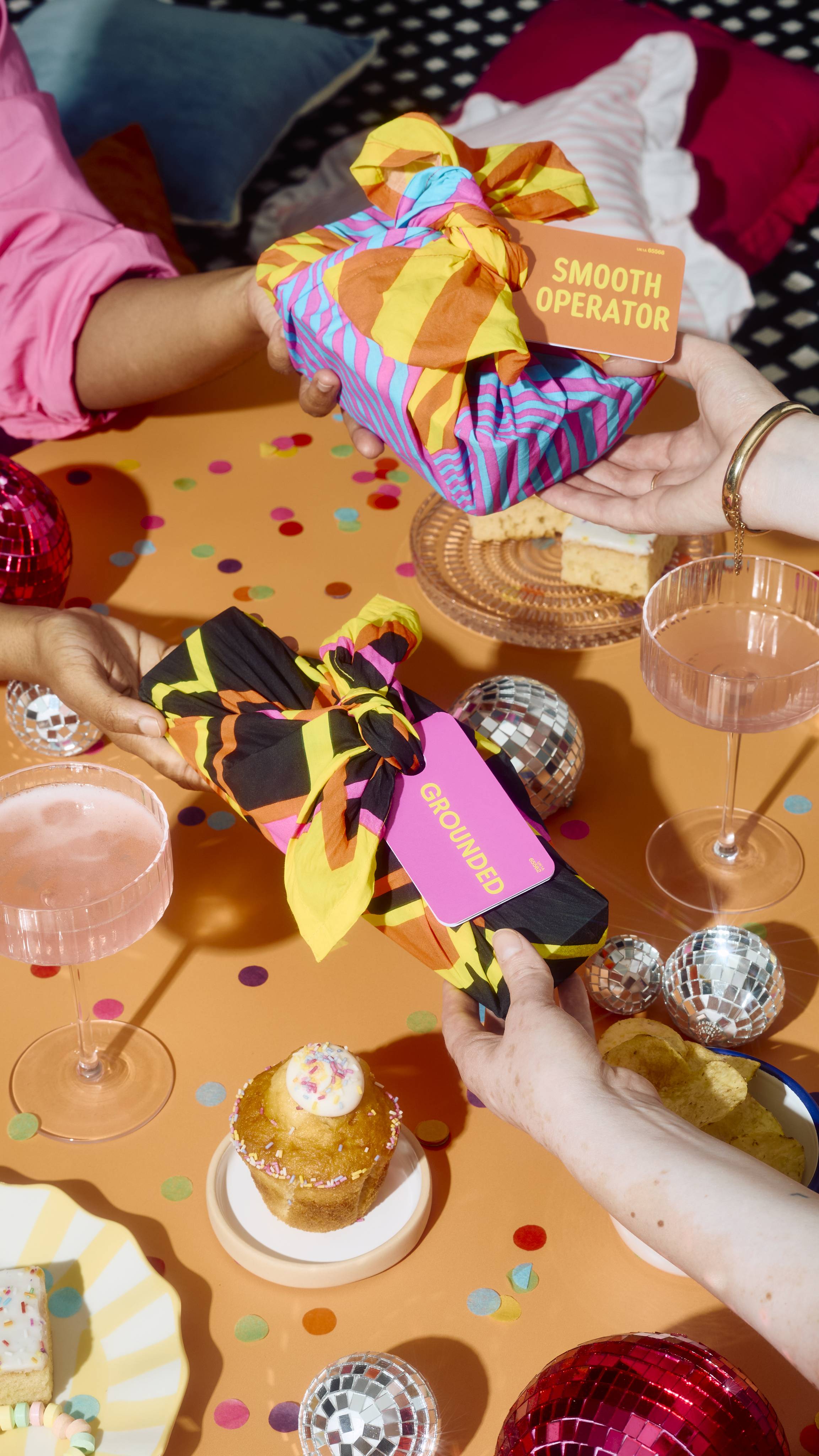 Hands pass knot-wrapped gifts tagged "SMOOTH OPERATOR" and "GROUNDED" across an orange party table decorated with mini disco balls, confetti, cupcakes, and drinks.