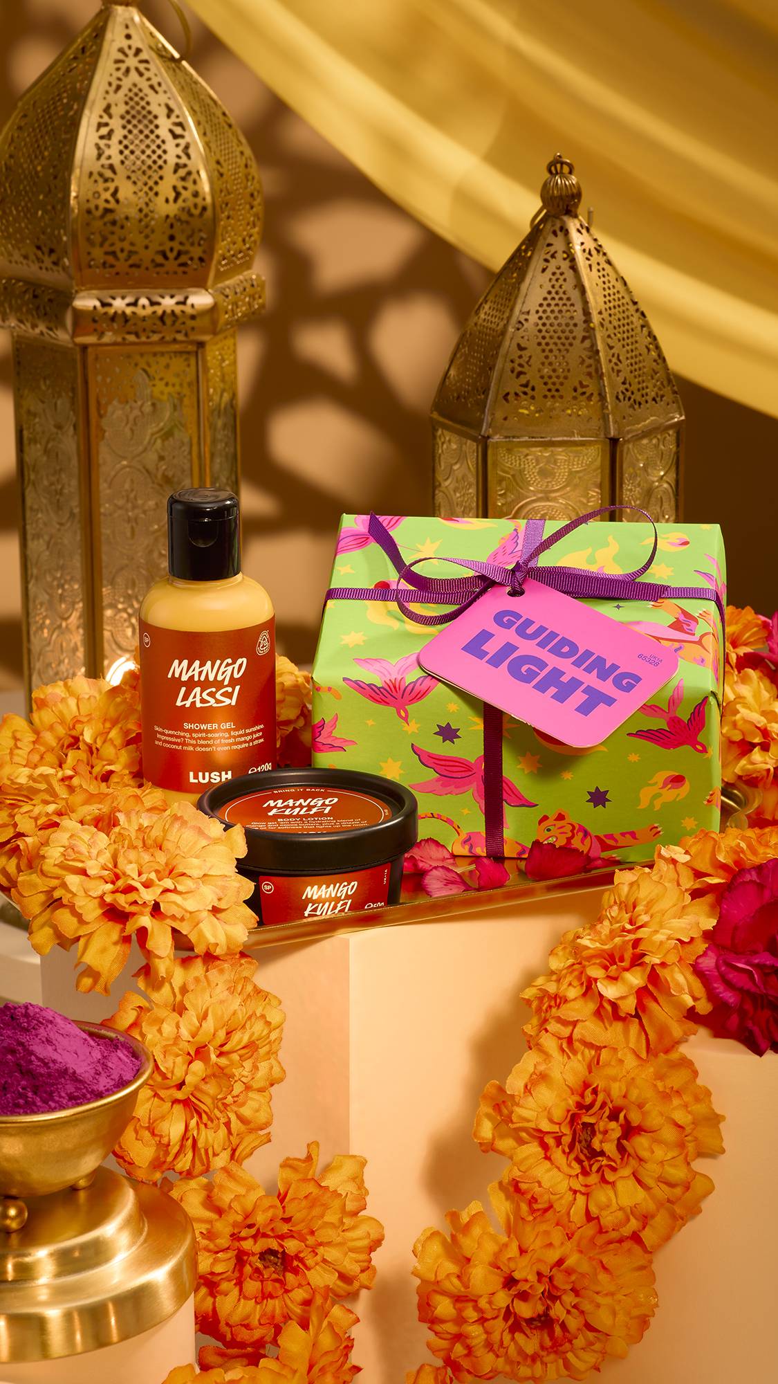A festive display of the Lush 'Guiding Light' gift set, featuring Mango Lassi shower gel, Mango Kulfi body lotion, and a green patterned gift box surrounded by marigold flowers, brass lanterns, and purple pigment in a warm Diwali setting.