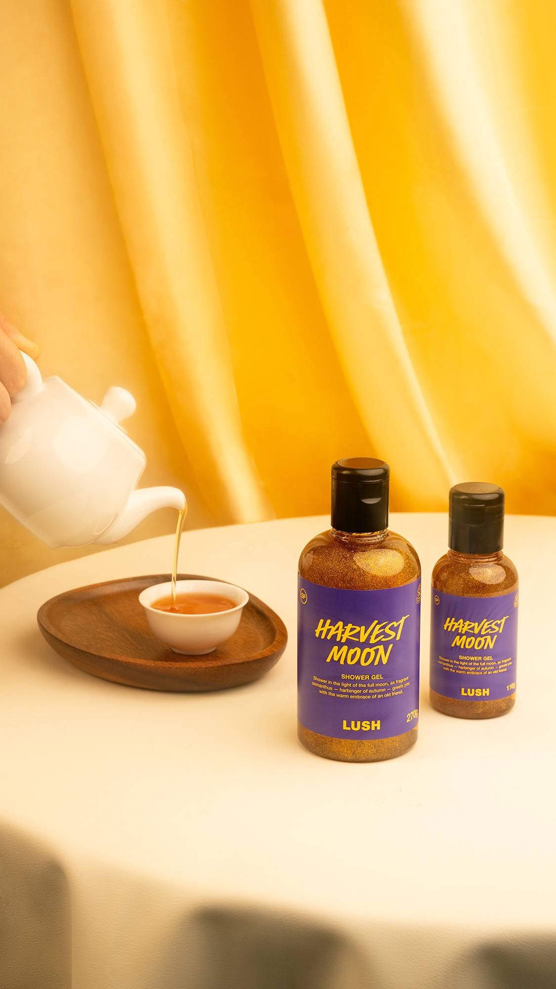 Two bottles of sparkly golden Lush "Harvest Moon" shower gel stand on a cream table. To the left, a hand pours tea from a white teapot into a small white teacup on a wooden tray, set against a draped gold backdrop.