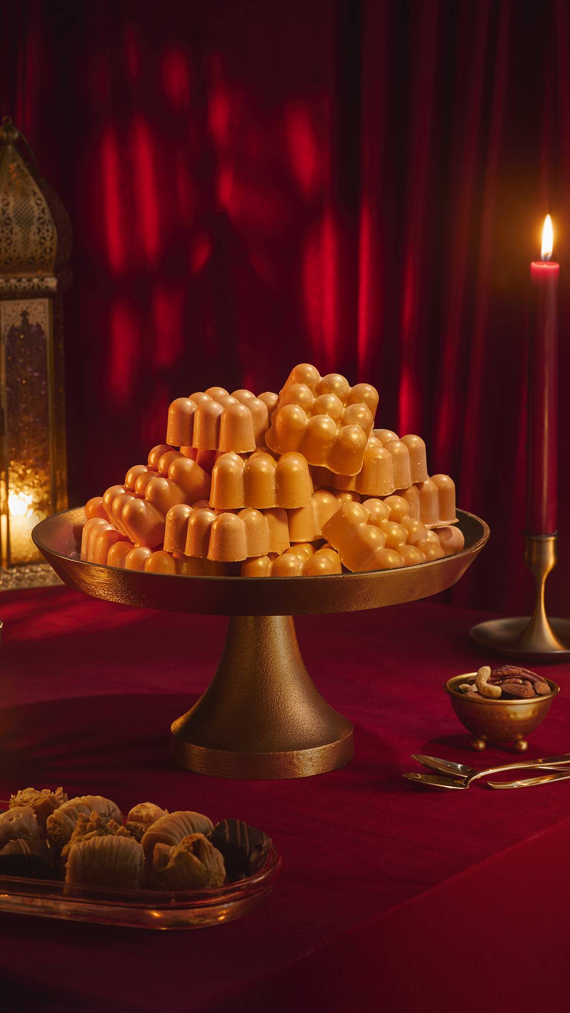 A large stack of shimmering, golden Hayl massage bars piled high on a brass cake stand. The festive setting features a red velvet tablecloth, a lit red candle, a bowl of nuts, and baklava, all set against a backdrop of draped red fabric.