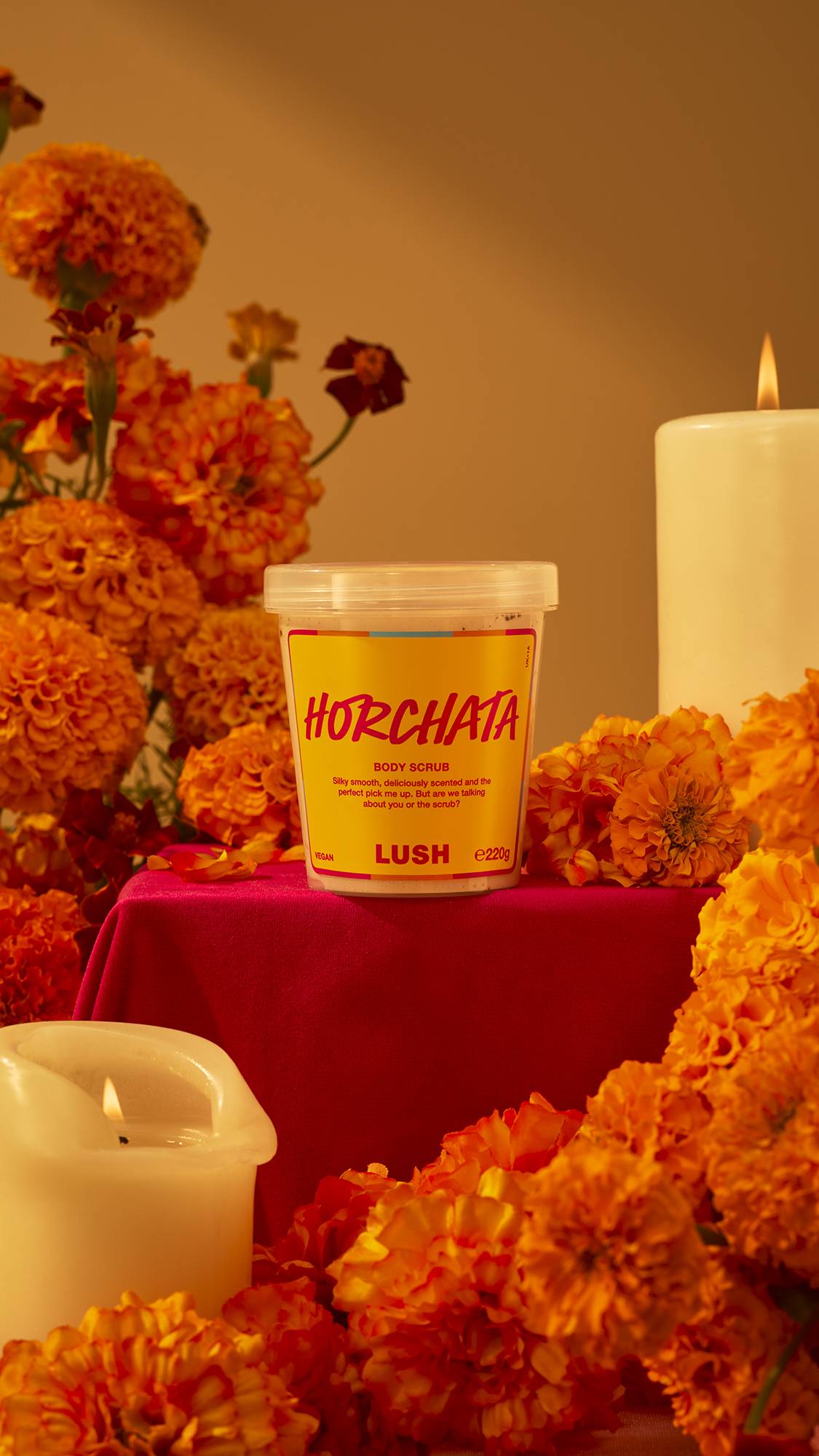 A pot of Lush Horchata body scrub with a yellow label sits on a red draped pedestal, surrounded by vibrant orange marigolds and lit candles in a warm, festive display.