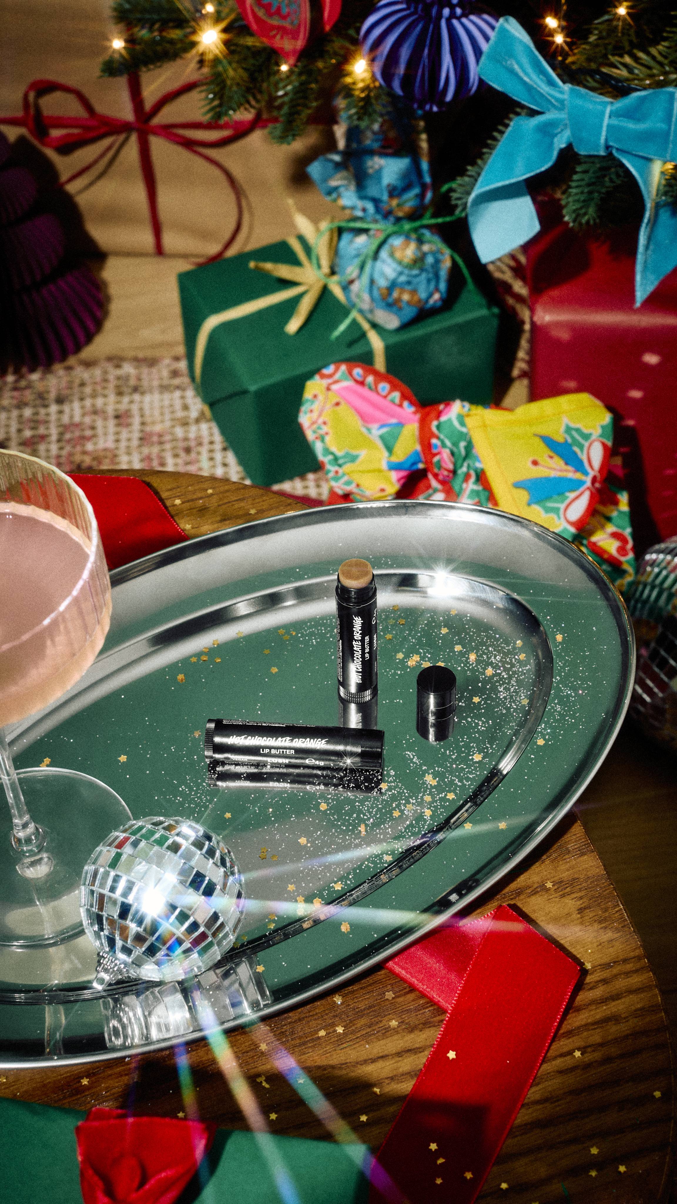 An open black tube of Lush Hot Chocolate Orange lip butter and its cap rest on a silver tray with gold glitter, alongside a cocktail glass and mini disco ball, against a backdrop of festive gifts.