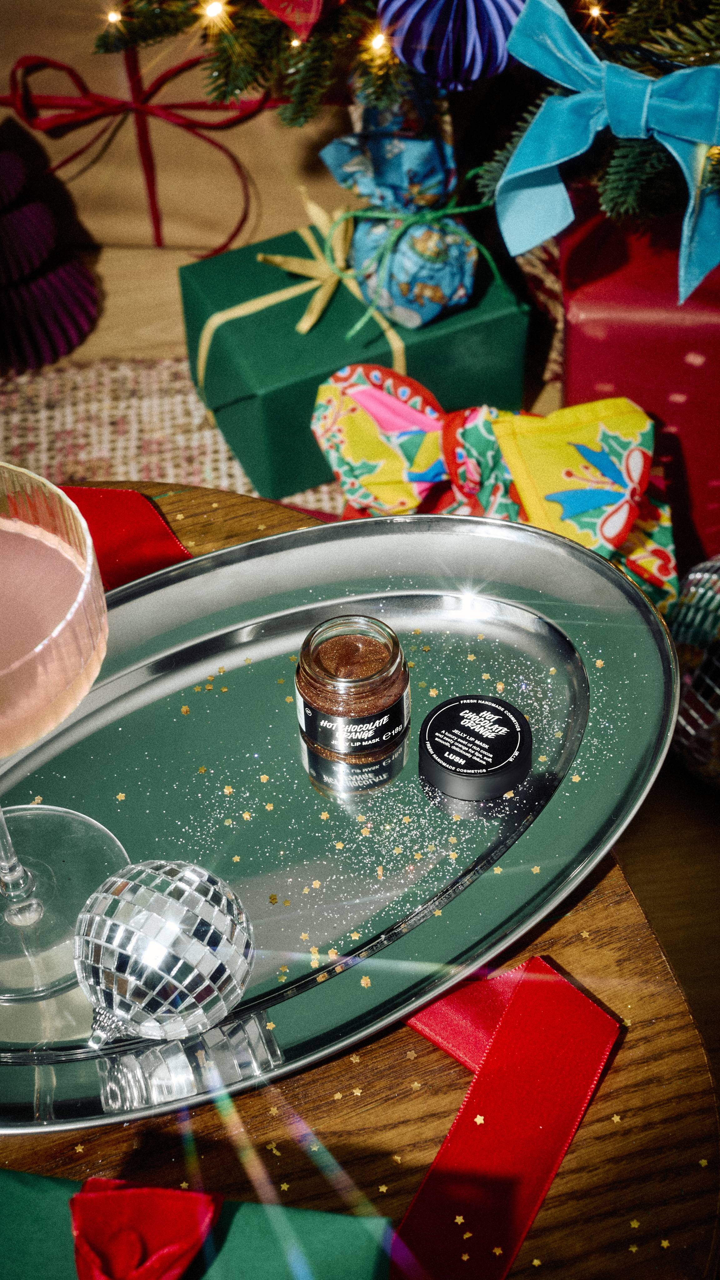 An open glass jar of Lush Hot Chocolate Orange lip mask and its lid rest on a silver tray scattered with gold glitter, alongside a cocktail glass and mini disco ball, with festive presents in the background.