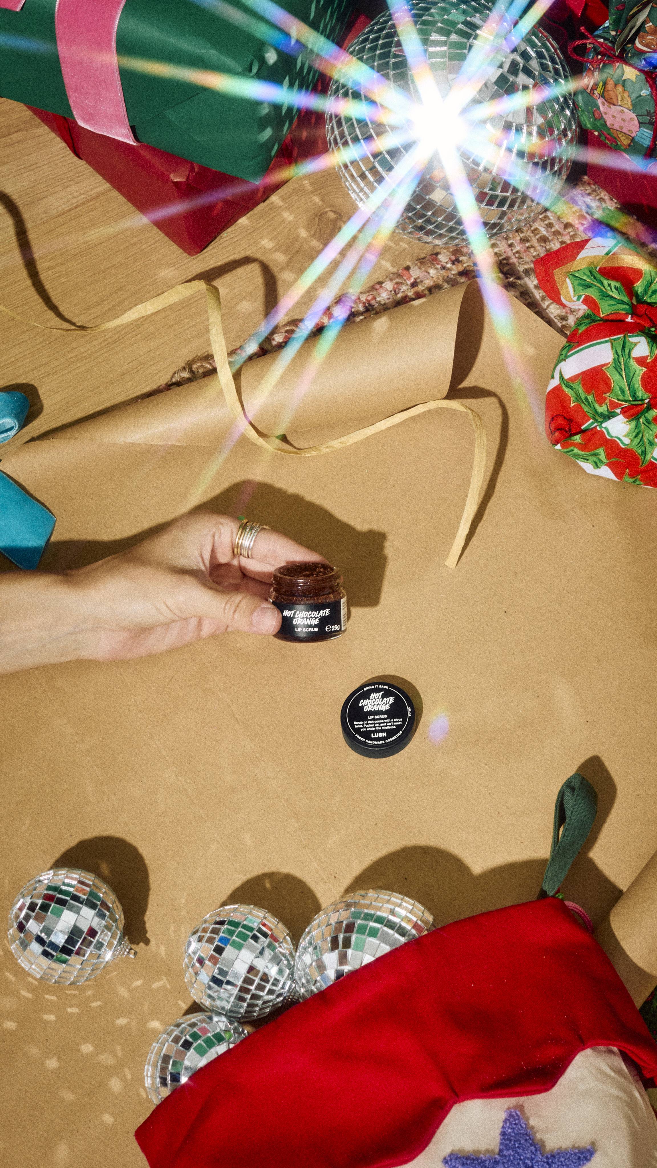 A hand holds an open glass jar of Lush Hot Chocolate Orange lip scrub on brown wrapping paper, surrounded by festive gifts, a disco ball, and a stocking filled with mirrored baubles.