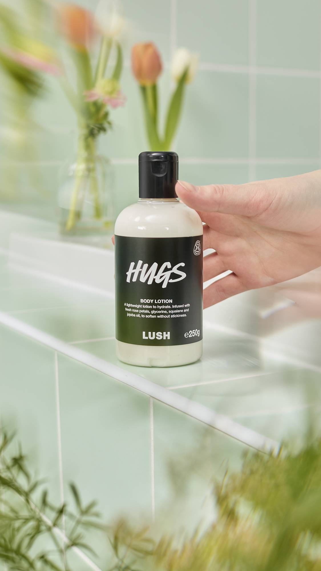 A person’s hand rests beside a bottle of Lush "Hugs" Body Lotion on a pale green tiled surface. The 250g bottle features a black label. In the soft-focus background are pink tulips and greenery, creating a fresh, botanical aesthetic.