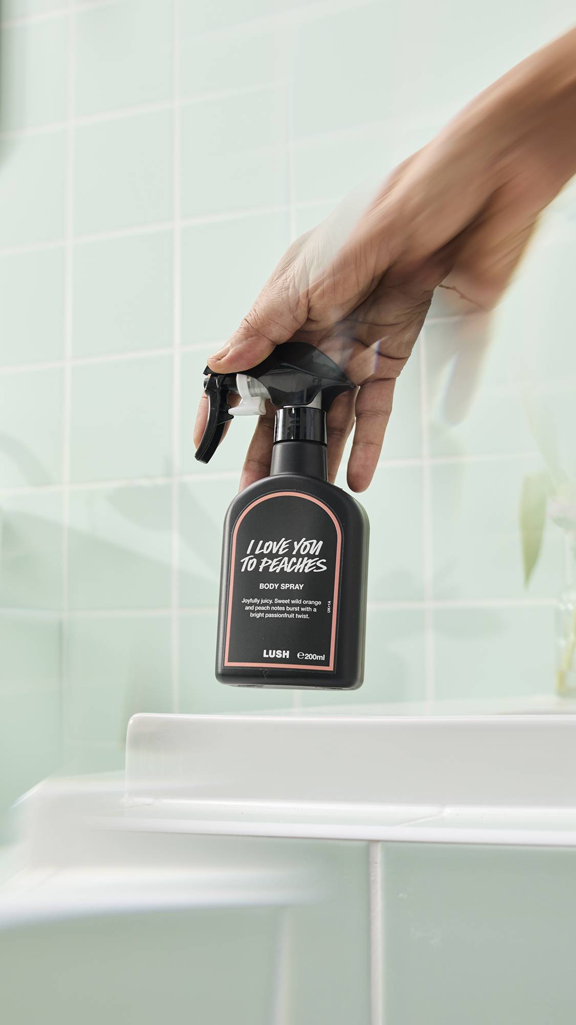A hand holds a black bottle of Lush "I Love You To Peaches" Body Spray in a brightly lit bathroom with pale green tiles. The hand is blurred as if moving the spray, with the product name and description clearly visible on the black label.