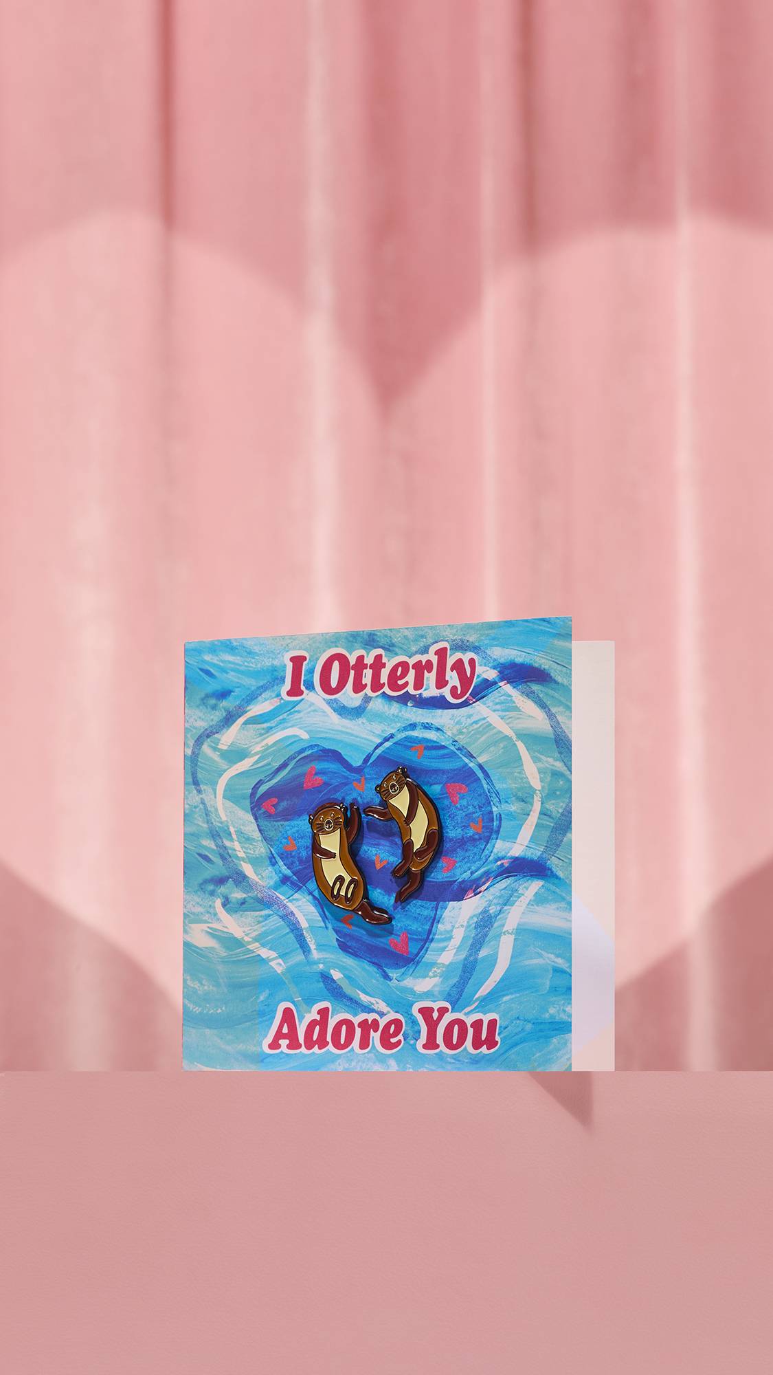 The "I Otterly Adore You" card features a blue swirling water design with the affectionate phrase in pink. Two brown otter enamel pins are attached to the front, floating together among small red hearts against a pink curtain background.