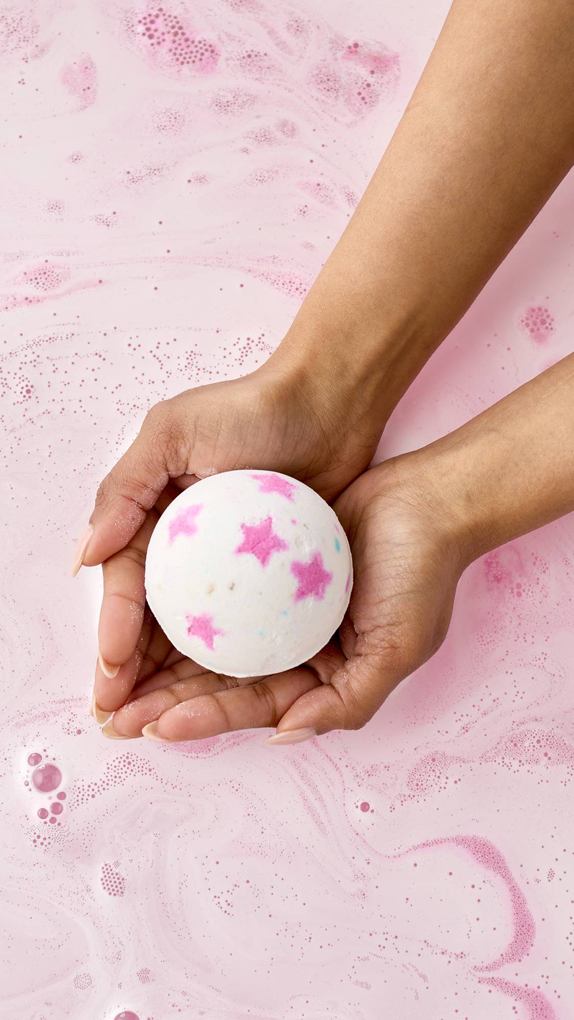 The dry, faceted Inclusion bath bomb sits on a plain background. The geometric product features crisp, color-blocked sections of bright pink, orange, and yellow.