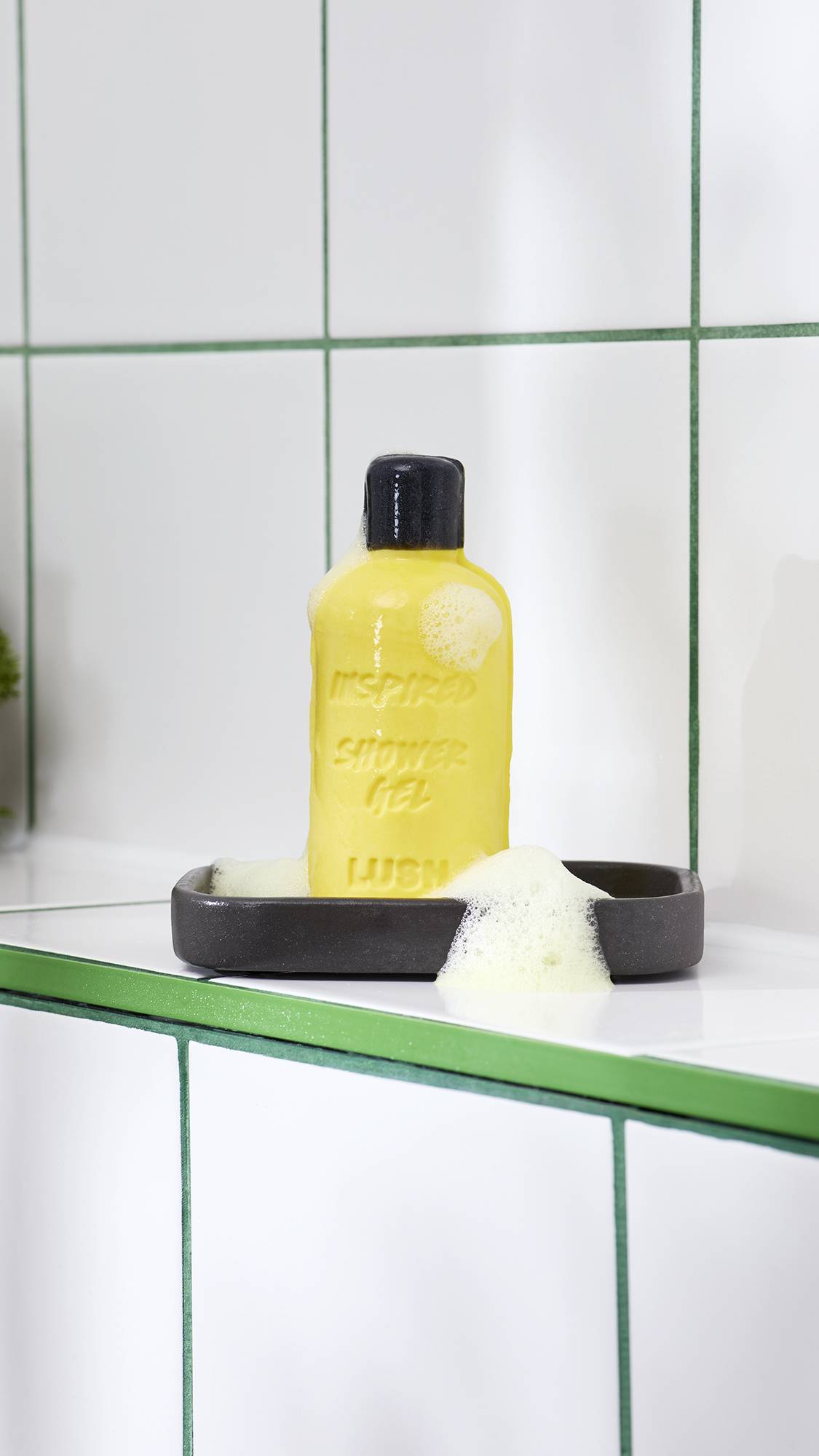 A bright yellow solid shower gel bar shaped like a bottle rests on a dark grey soap dish, covered in light, pale yellow foam. It sits against a backdrop of white tiled walls with green grouting.