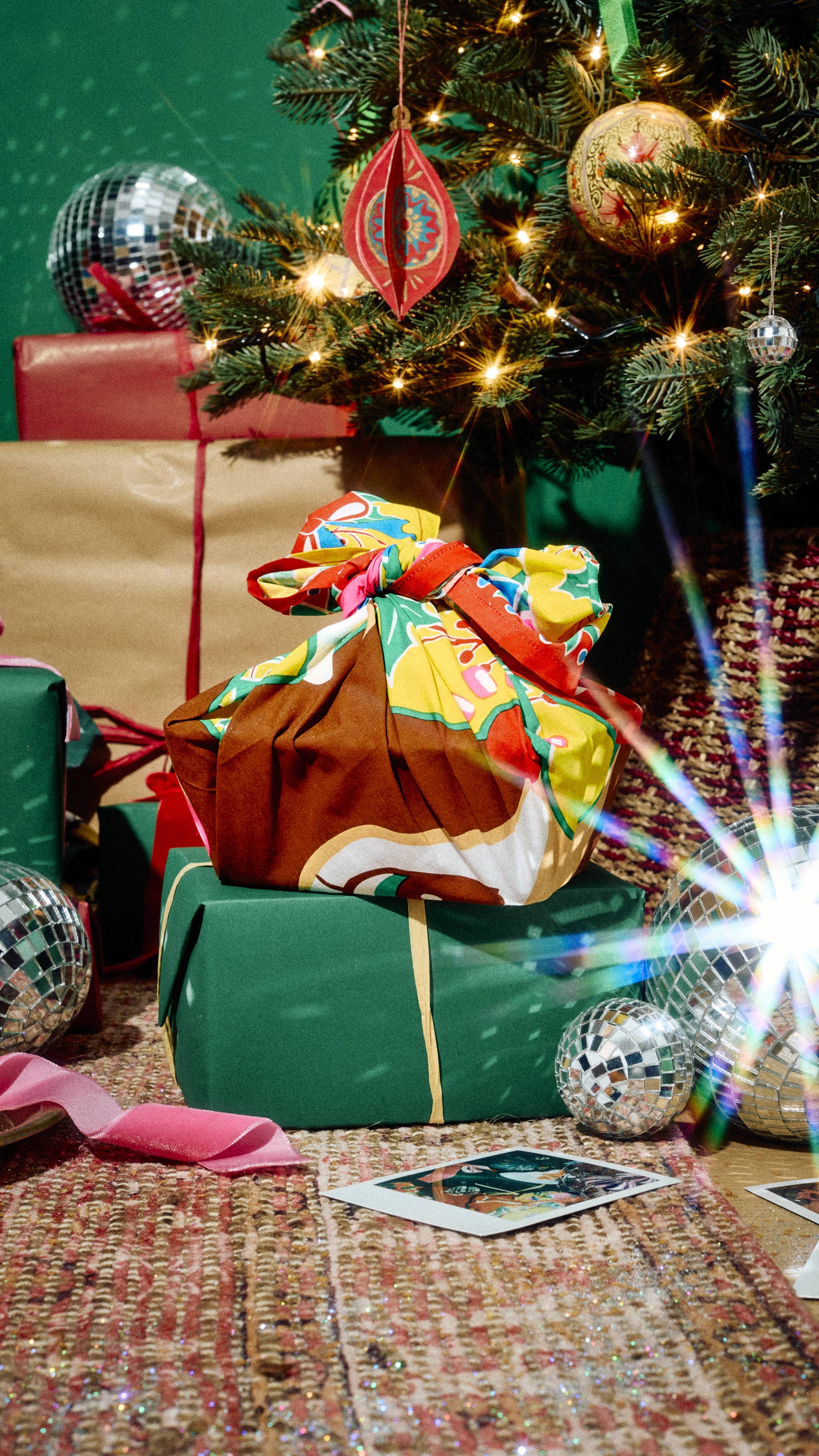 A gift wrapped in a colourful reindeer-patterned Lush fabric Knot Wrap sits atop a green gift box, displayed on a rug near a decorated Christmas tree with sparkly disco balls.