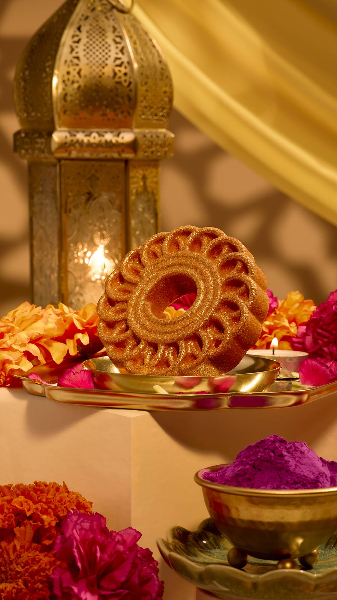 A circular, shimmery golden Lush bath bomb with a carved wheel design sits on a gold dish, surrounded by orange marigolds, pink petals, lit tealights, a brass lantern, and purple powder pigment in a warm Diwali setting.