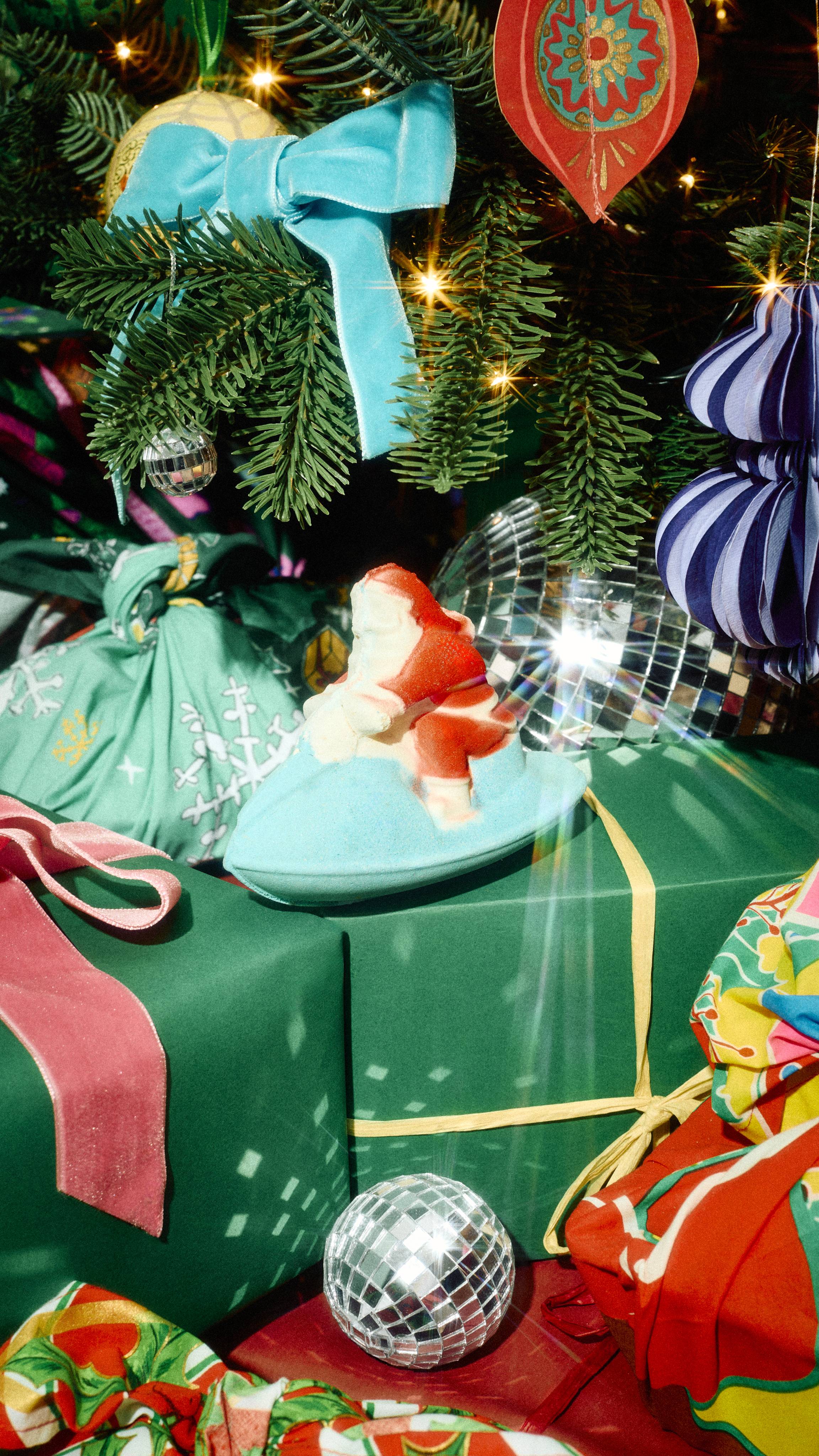 A Santa on a jet ski bath bomb sits on a green gift box beneath a Christmas tree decorated with mini disco balls, velvet bows, and wrapped presents.