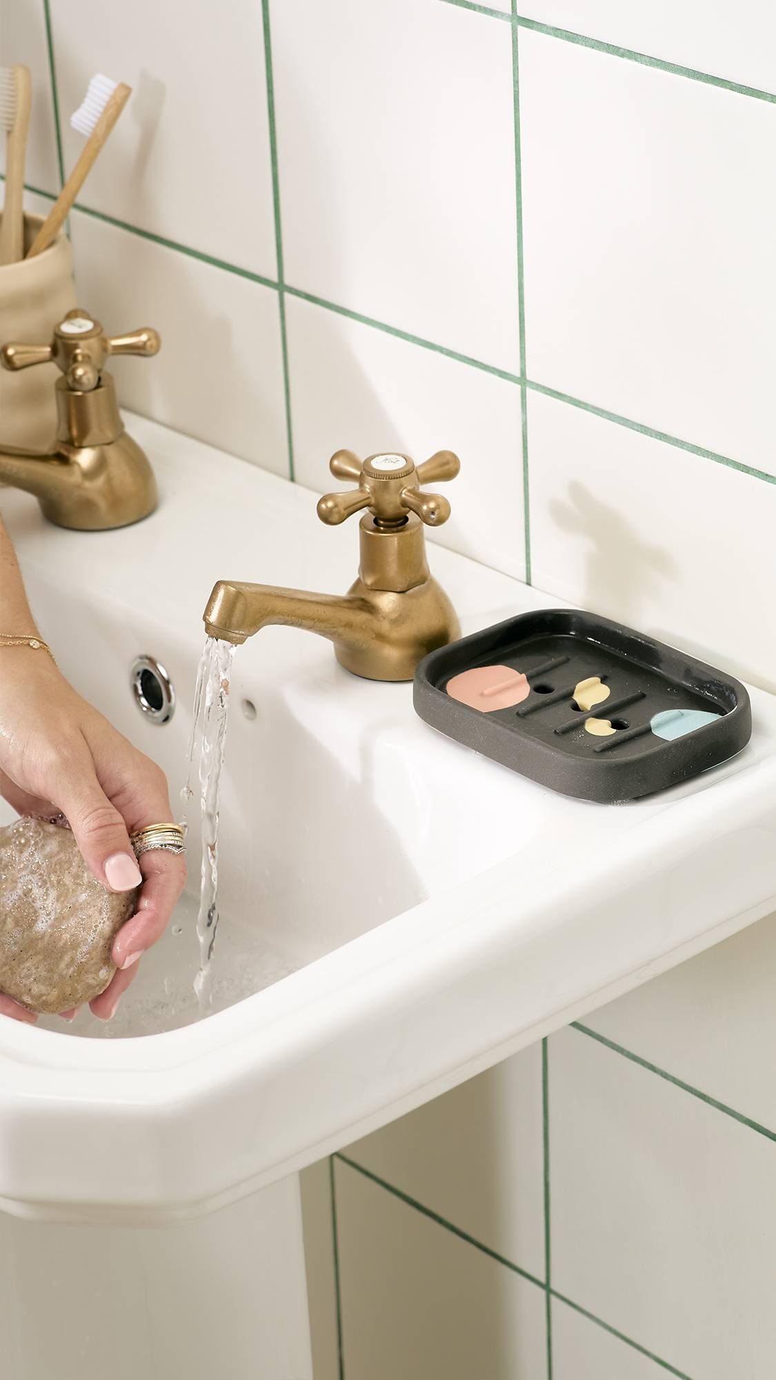 A person holds a bar of soap under running water from a brass tap over a white bathroom sink, with a dark grey ceramic soap dish featuring pastel spots resting beside the tap against white tiled walls.