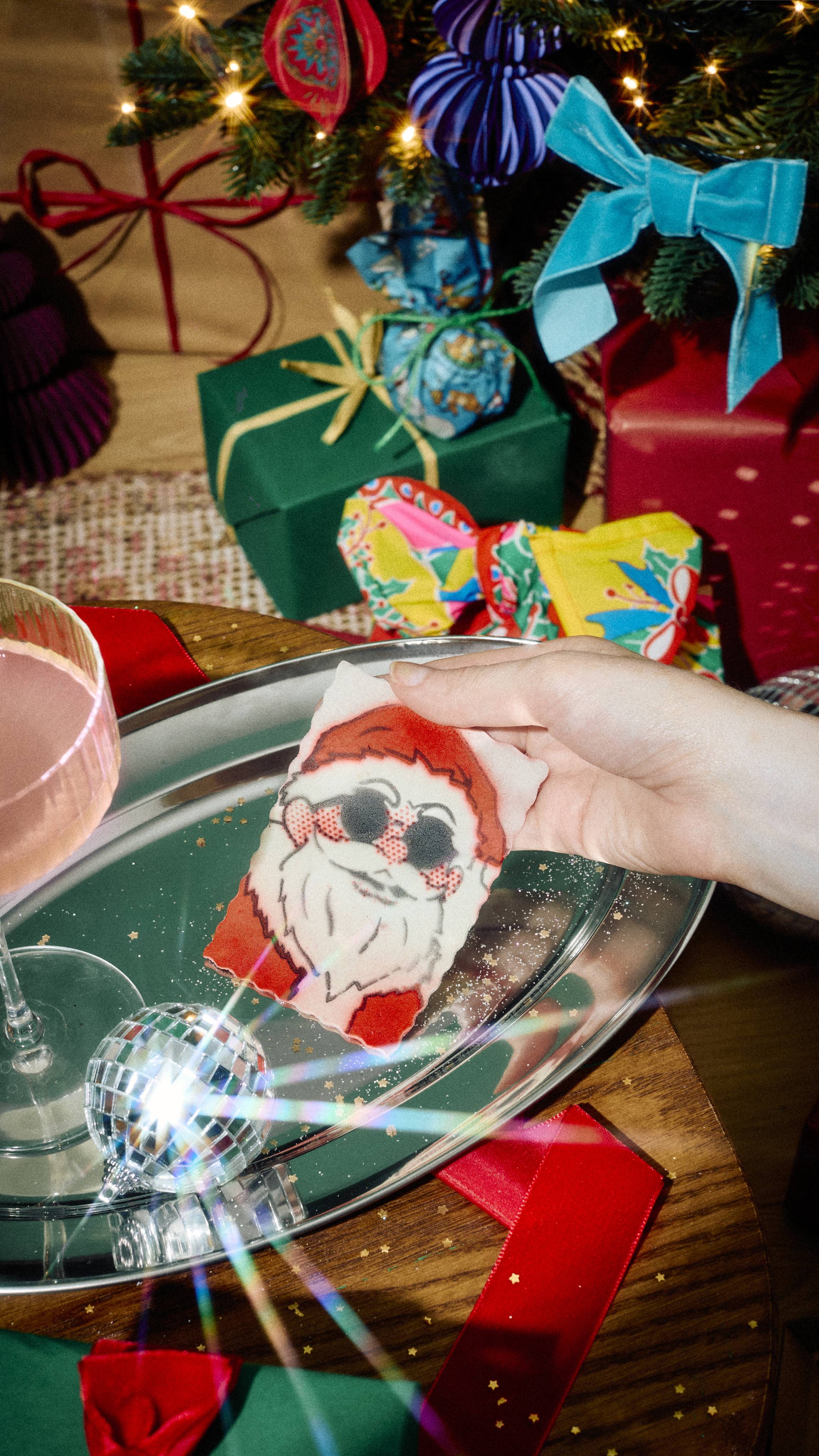 A hand holds a large stamp-shaped sheet of surfactant paper featuring Santa Claus in sunglasses over a silver tray, set against a festive background with wrapped gifts and a decorated Christmas tree.