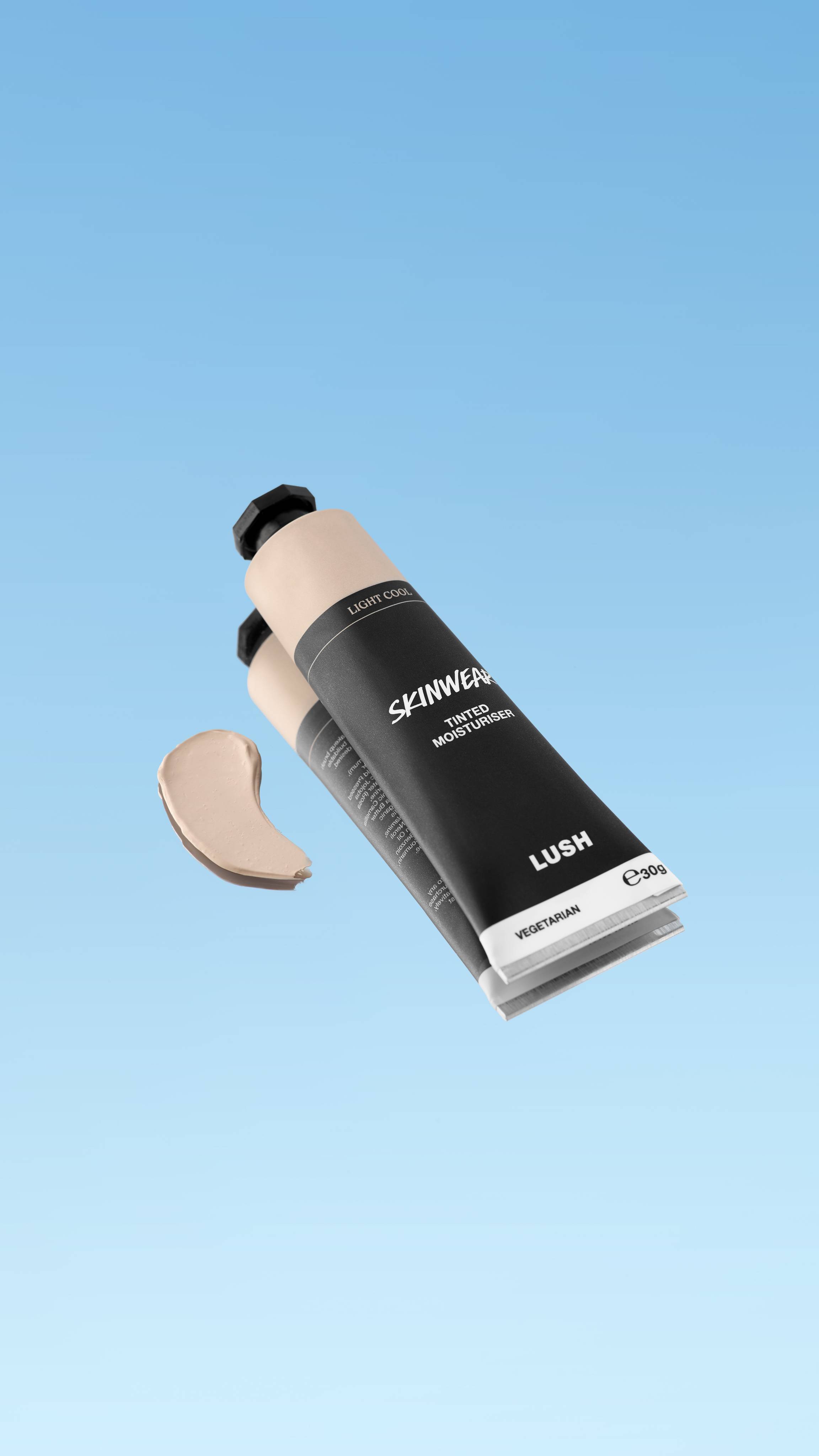An angled shot of a black and pale beige LUSH Skinwear Tinted Moisturiser tube in the shade Light Cool. A small swatch of the product floats next to the tube, all suspended against a bright, gradient blue background.