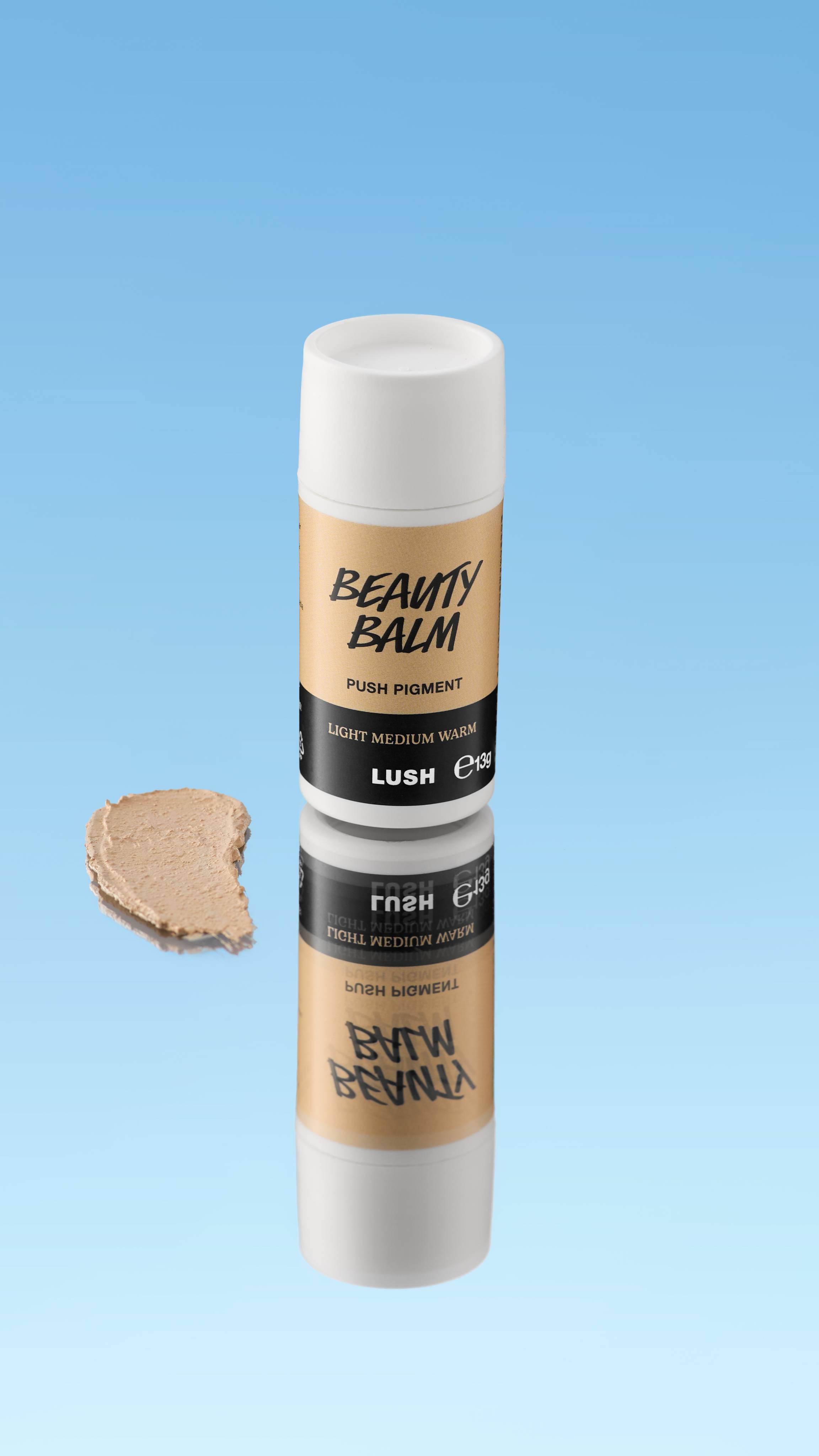 An angled product shot of a white and peach-beige LUSH Beauty Balm Push Pigment stick in the shade Light Medium Warm. A thick swatch of the warm beige product rests next to the reflected stick, all against a bright blue background.
