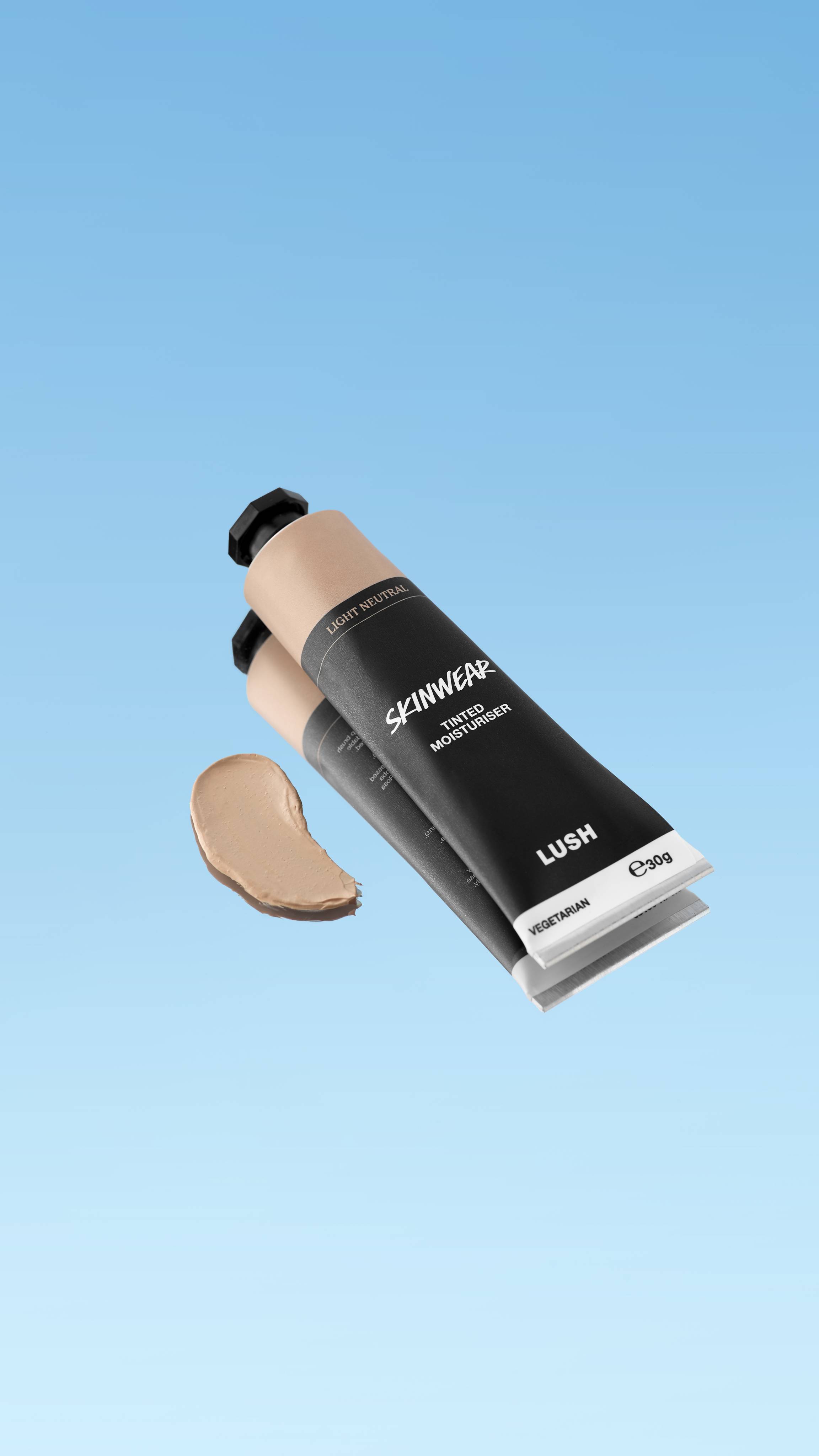 A product shot showing the black tube of Skinwear Tinted Moisturiser in the Light Neutral shade, floating against a blue gradient background. A dollop of pale, balanced beige cream is swatched to the left of the tube.