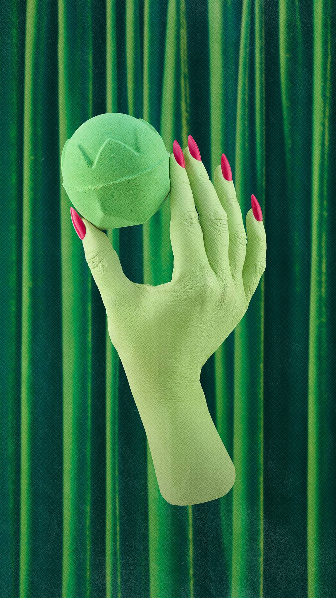 A light-green hand with bright pink fingernails holds up a matching light-green carved bath bomb, set against a backdrop of draped emerald-green velvet curtains.