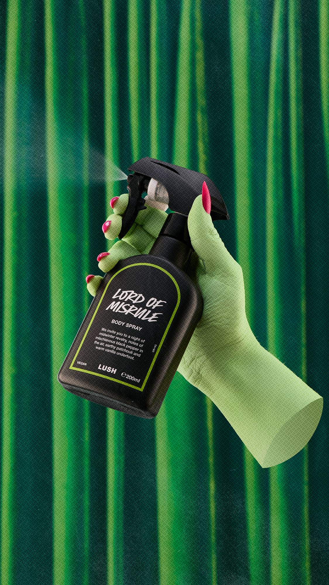 A green-skinned hand with hot pink nails sprays Lush Lord of Misrule body spray, creating a fine mist against a lush backdrop of dark green velvet drapes.