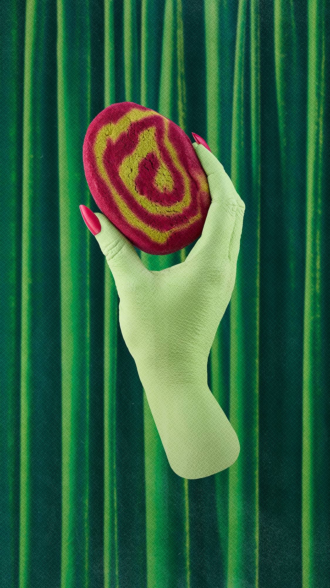A green hand with long pink nails holds a magenta and lime green swirled "Lord of Misrule" bubble bar, set against a dark green draped velvet curtain background.