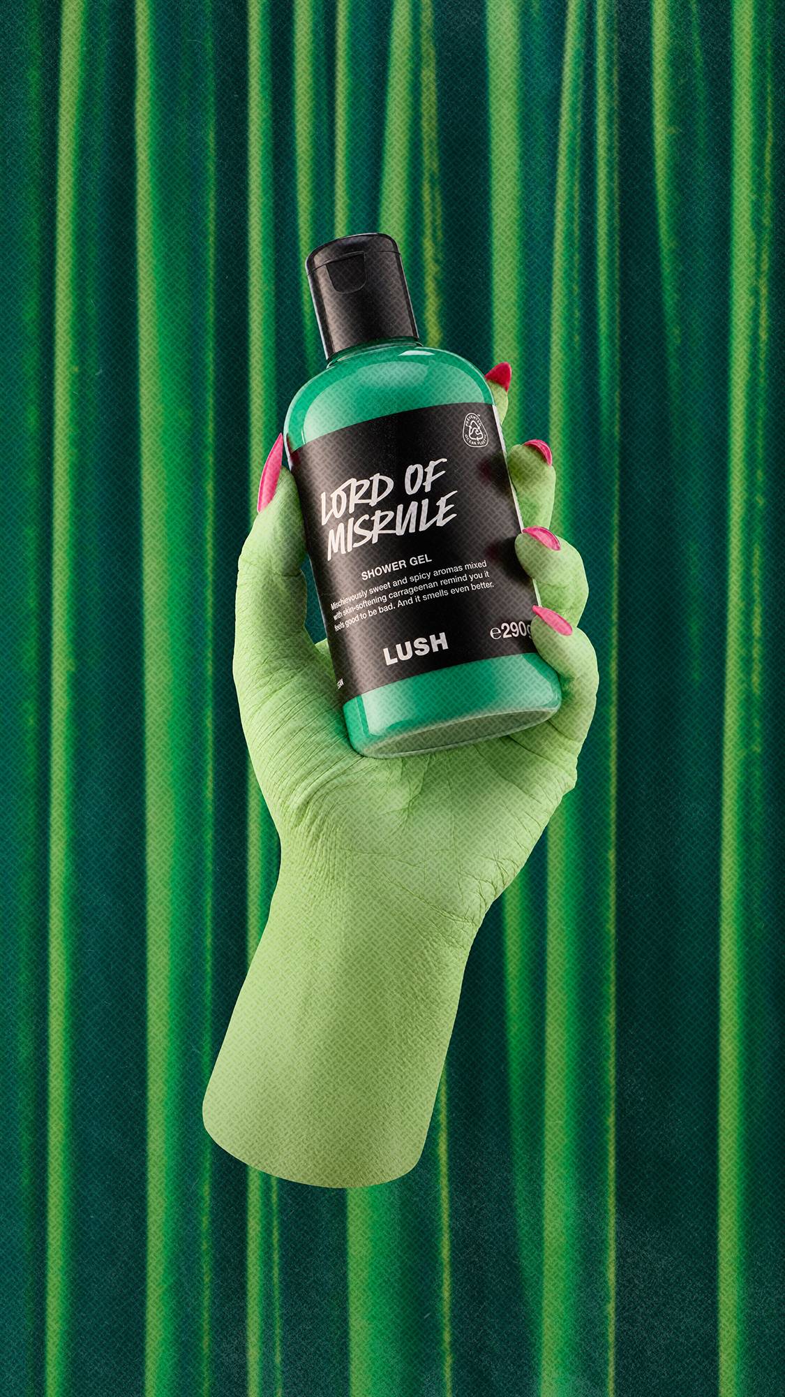 A green hand with bright pink nails holds a bottle of Lush Lord of Misrule shower gel, filled with vibrant green liquid, set against a dark green draped velvet curtain background.