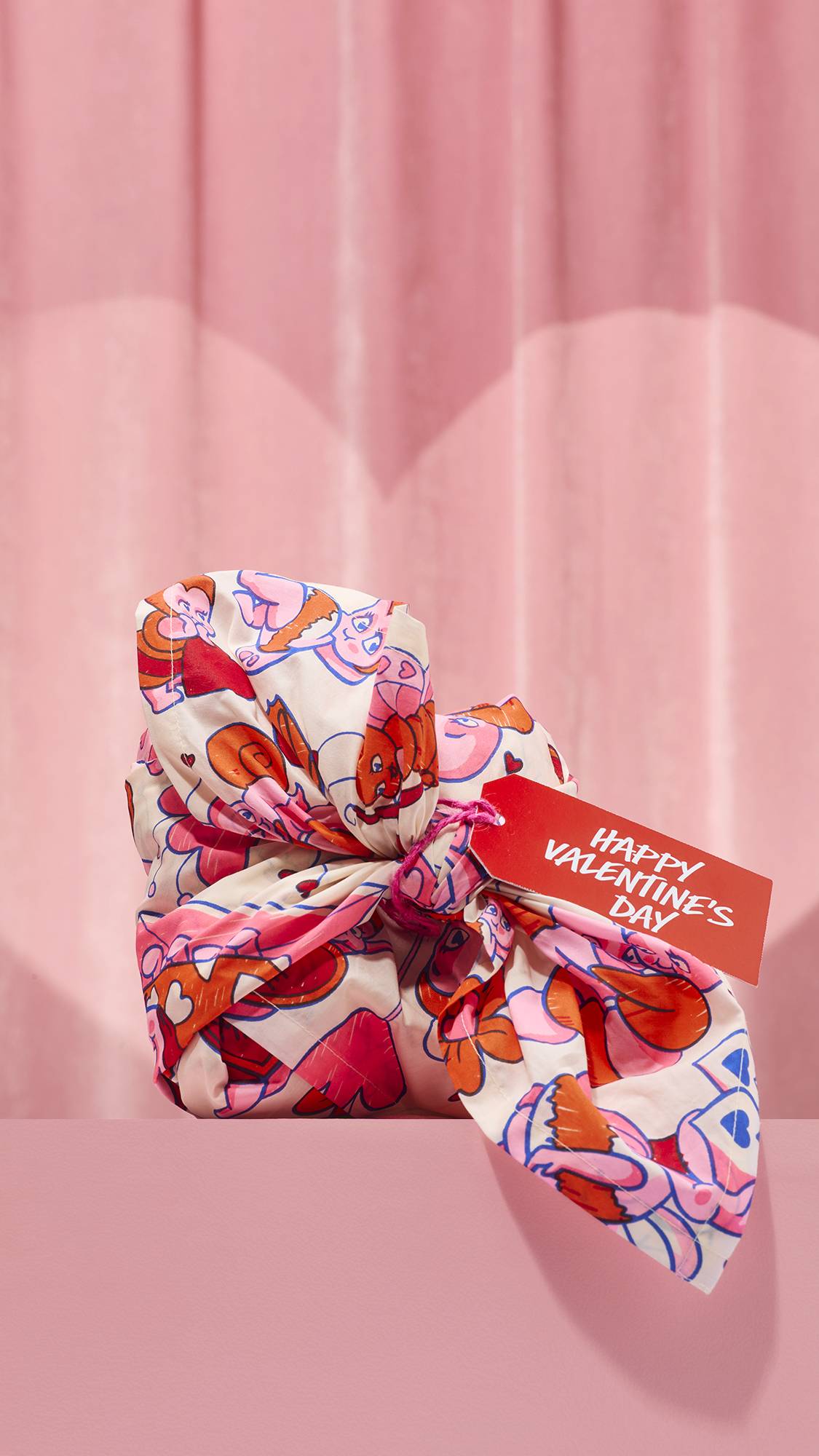 The Love Bug Knot Wrap features a white fabric tied around a gift, decorated with a playful pattern of pink and orange cartoon love bugs. A red tag reading "Happy Valentine's Day" hangs from the knot against a soft pink curtain background.