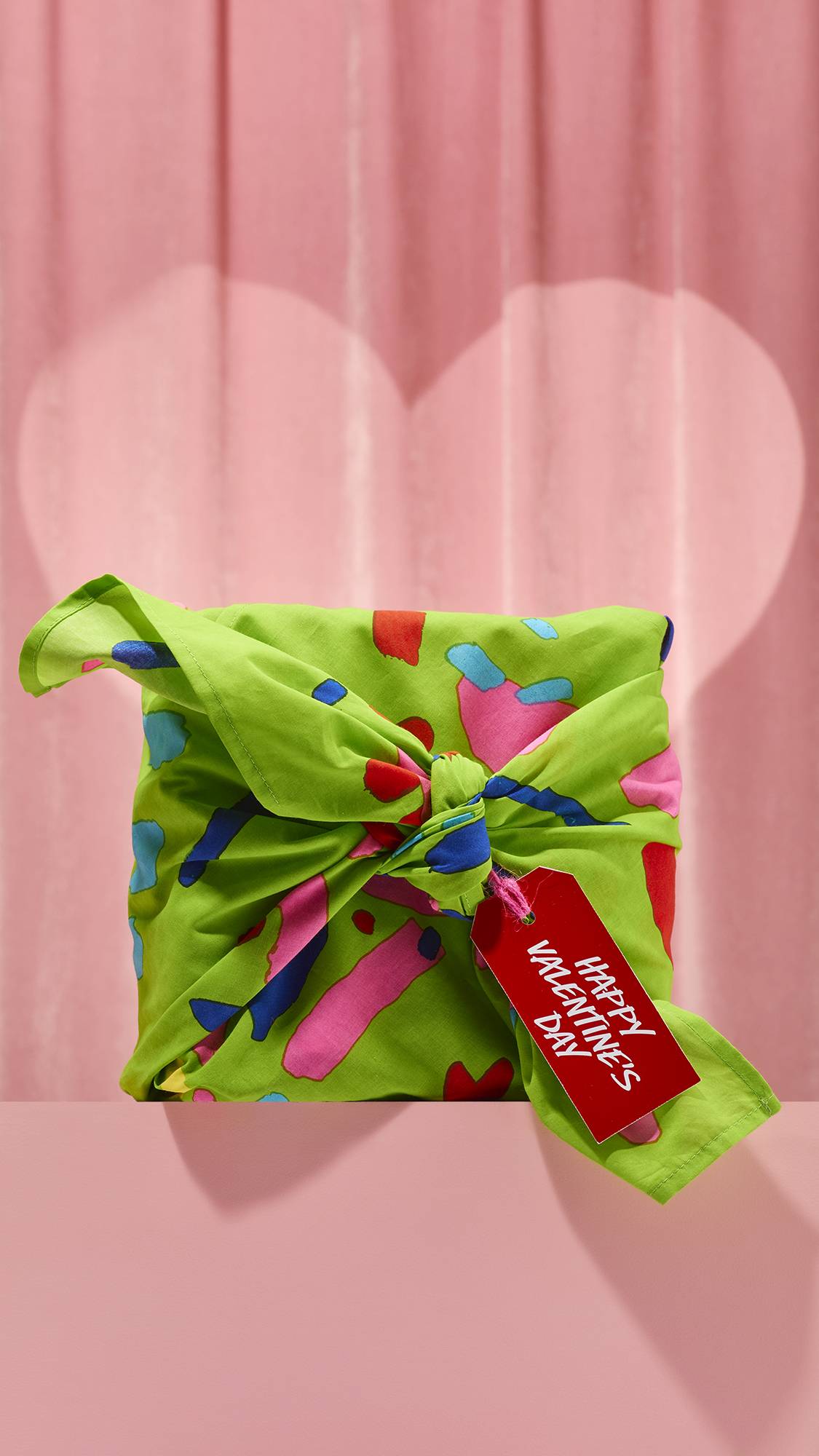 The bright green Love Knot Knot Wrap is tied around a gift, featuring abstract pink, blue, and red shapes. A red tag reading Happy Valentine's Day hangs from the knot against a soft pink background.