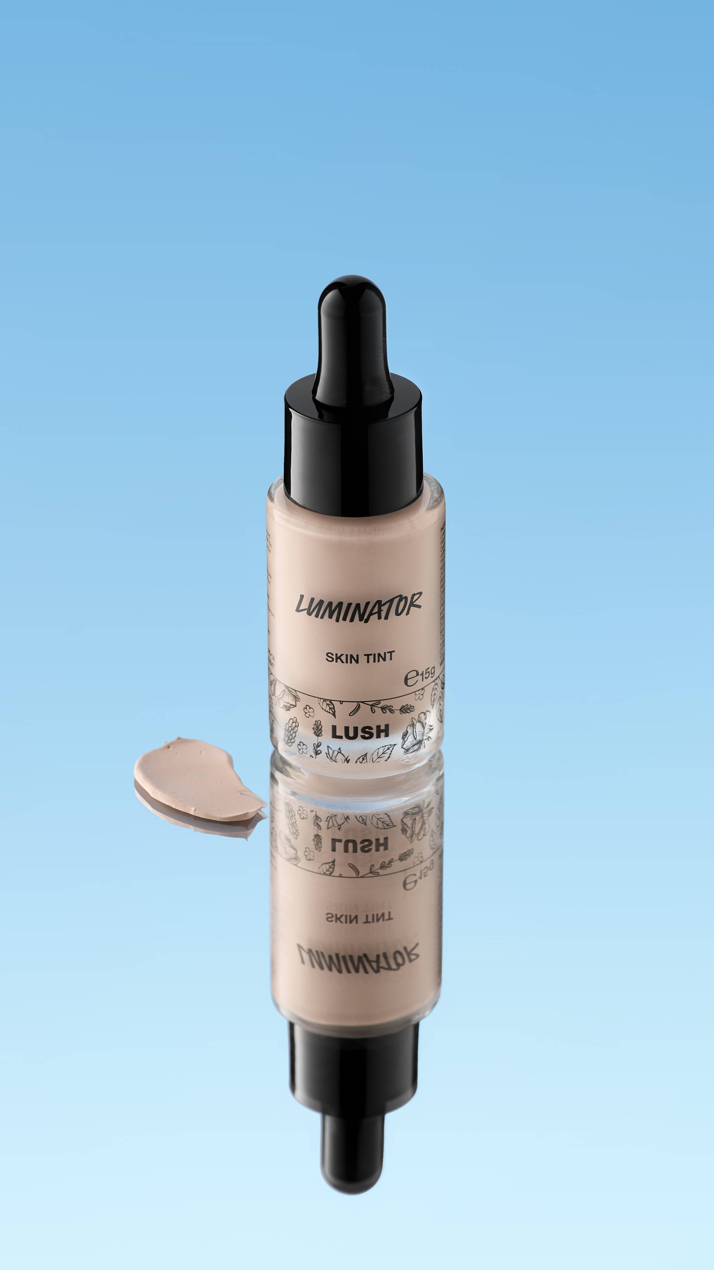 
An angled product shot of a dropper bottle of LUSH Luminator Skin Tint with a black cap, standing on a reflective surface. A creamy rosy-beige swatch of the product rests next to the bottle and its reflection, all against a bright blue background.