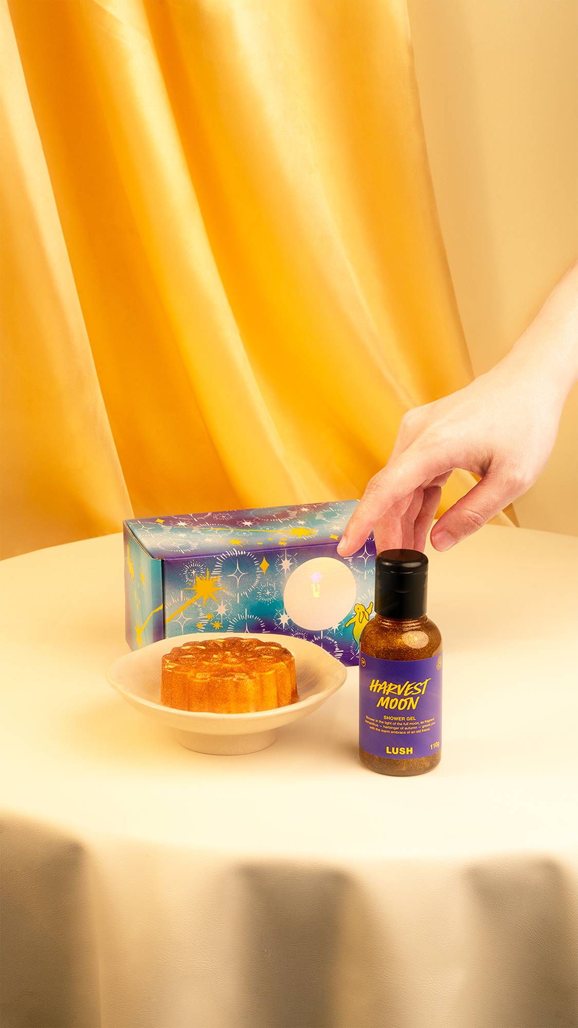 A hand reaches towards a small bottle of glittery golden Lush "Harvest Moon" shower gel on a cream tablecloth. A starry purple gift box and a mooncake bath bomb on a small white plate sit behind it, against a draped gold backdrop.