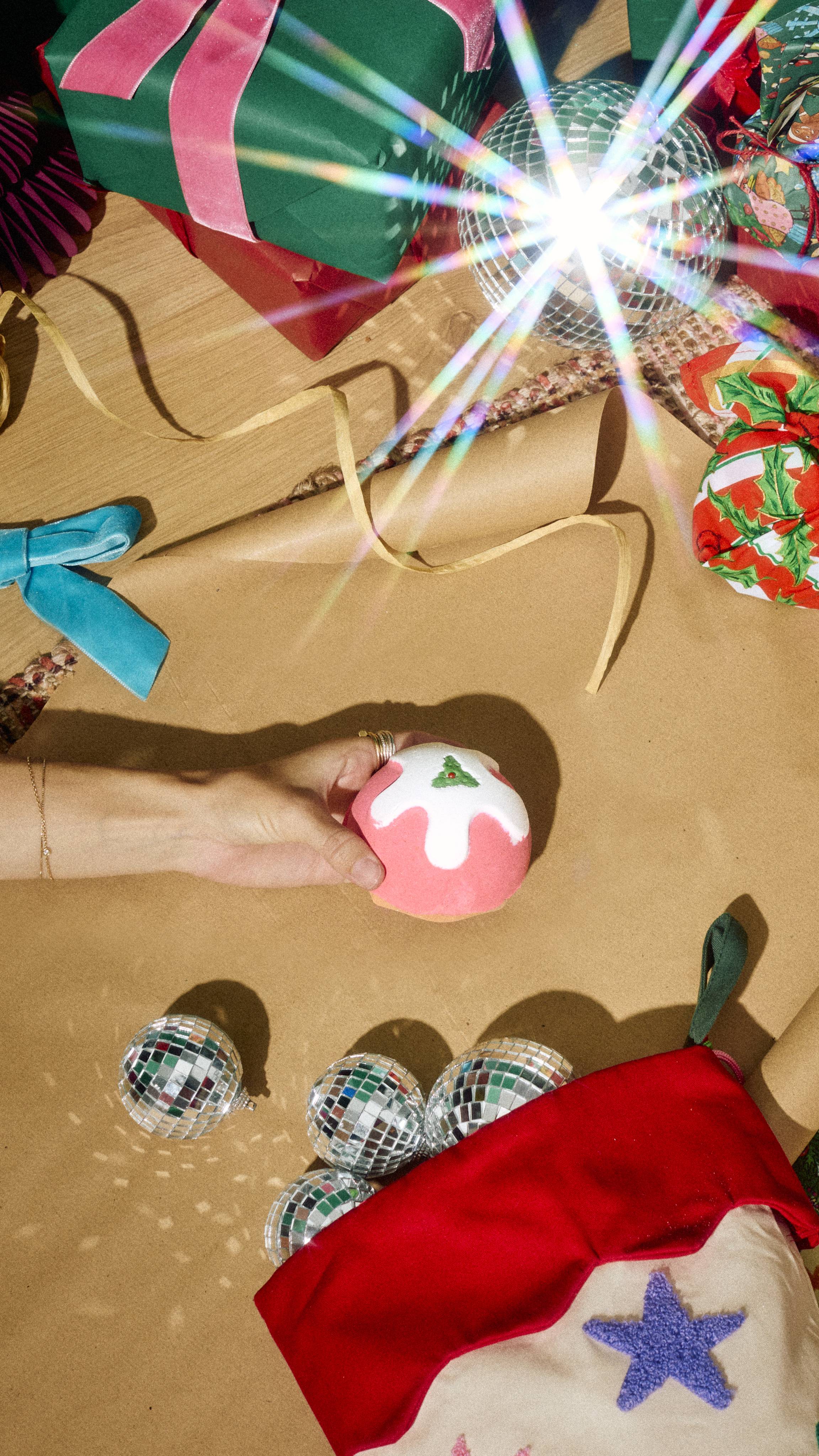 A hand reaches for a Christmas pudding bath bomb on a kraft paper workspace, surrounded by wrapped gifts, disco balls, ribbons, and a red Christmas stocking spilling mirrored baubles.