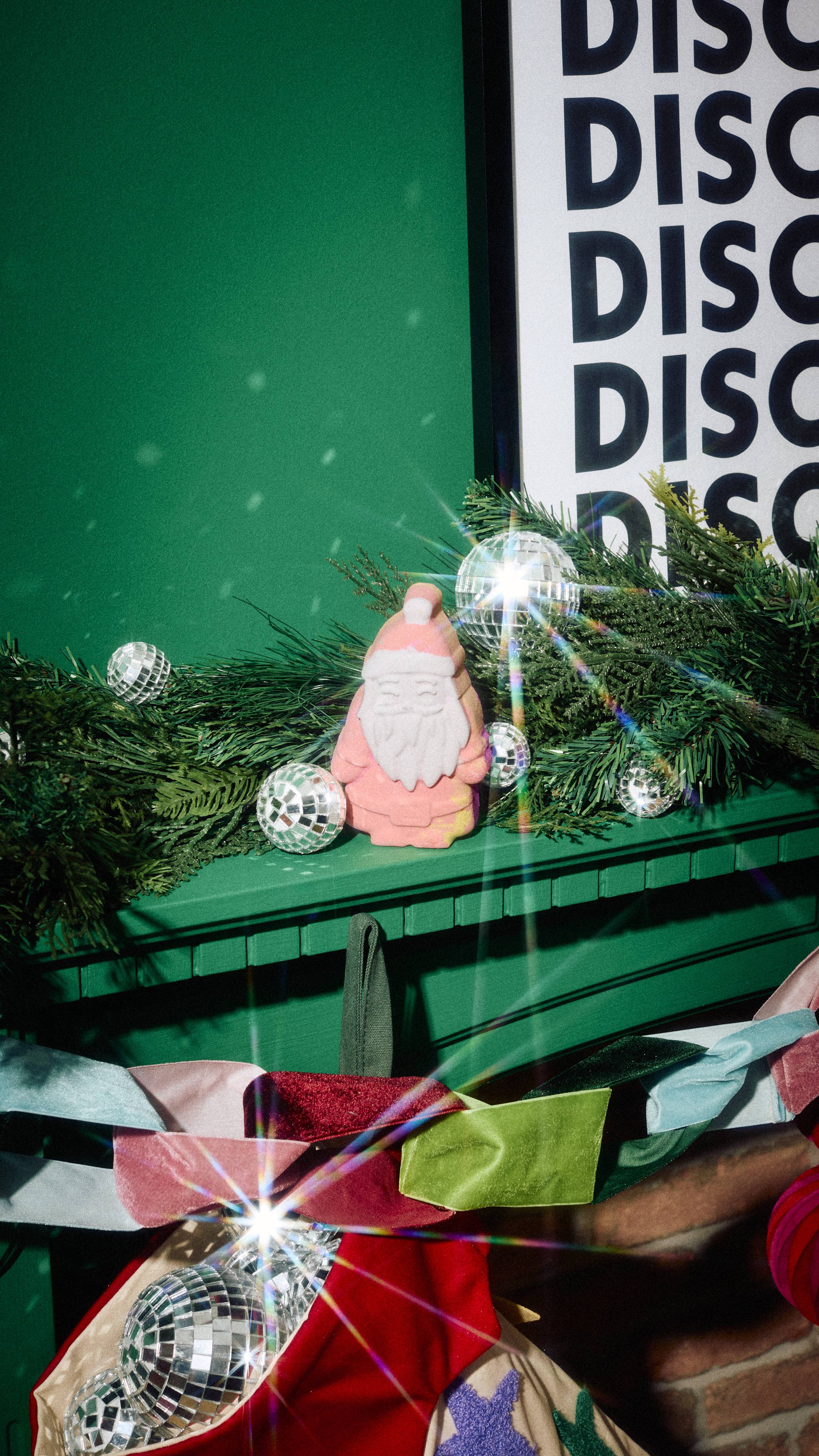 A pink Lush "Magical Santa" bath bomb sits on a green mantelpiece nestled among pine garland and mini disco balls, with a colourful velvet bow garland and stocking below.