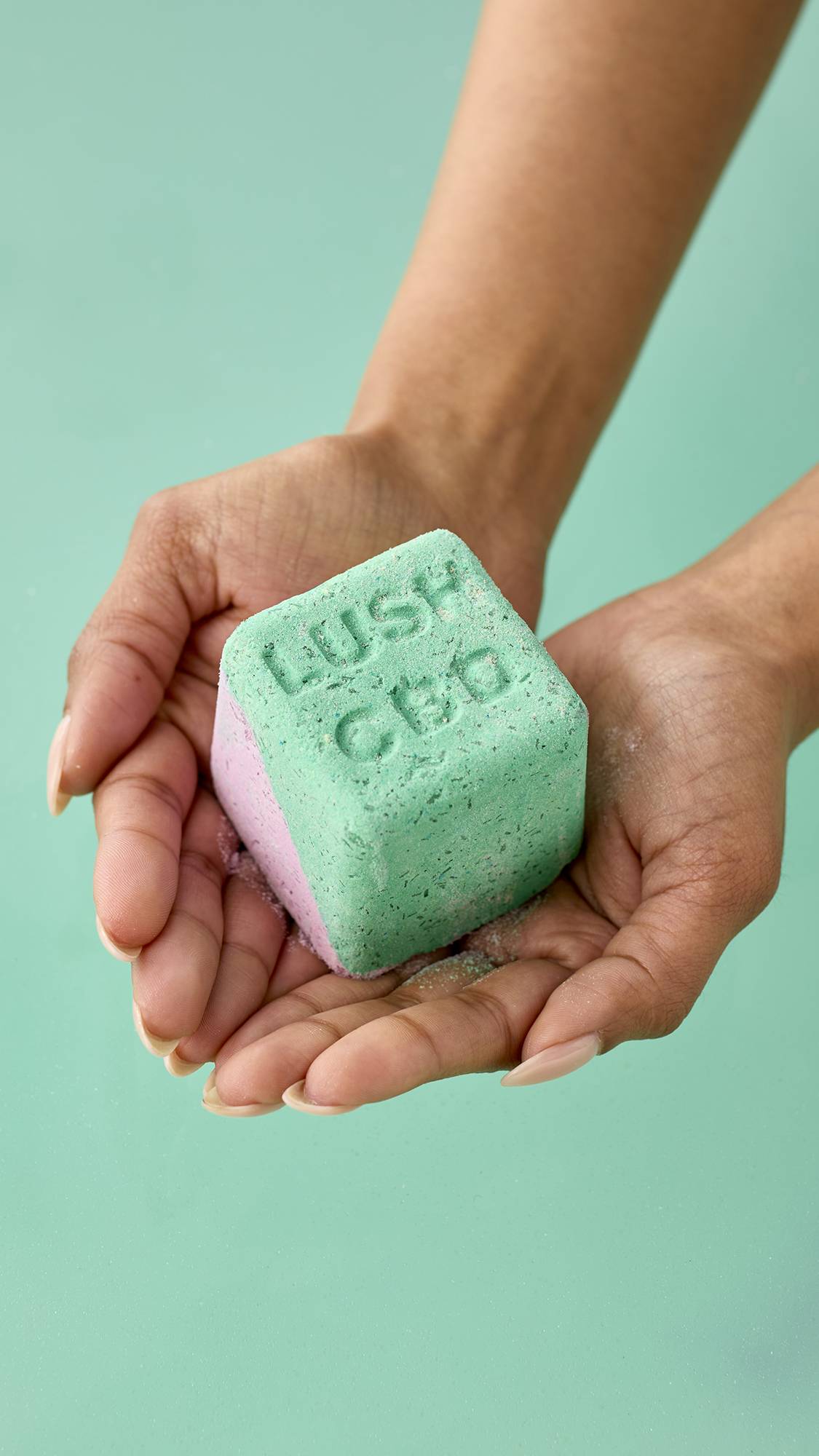 A pair of hands holds a cube-shaped bath bomb with a split green and purple design. The top green face has "LUSH CBD" embossed into the surface. It is held against a solid mint green background.