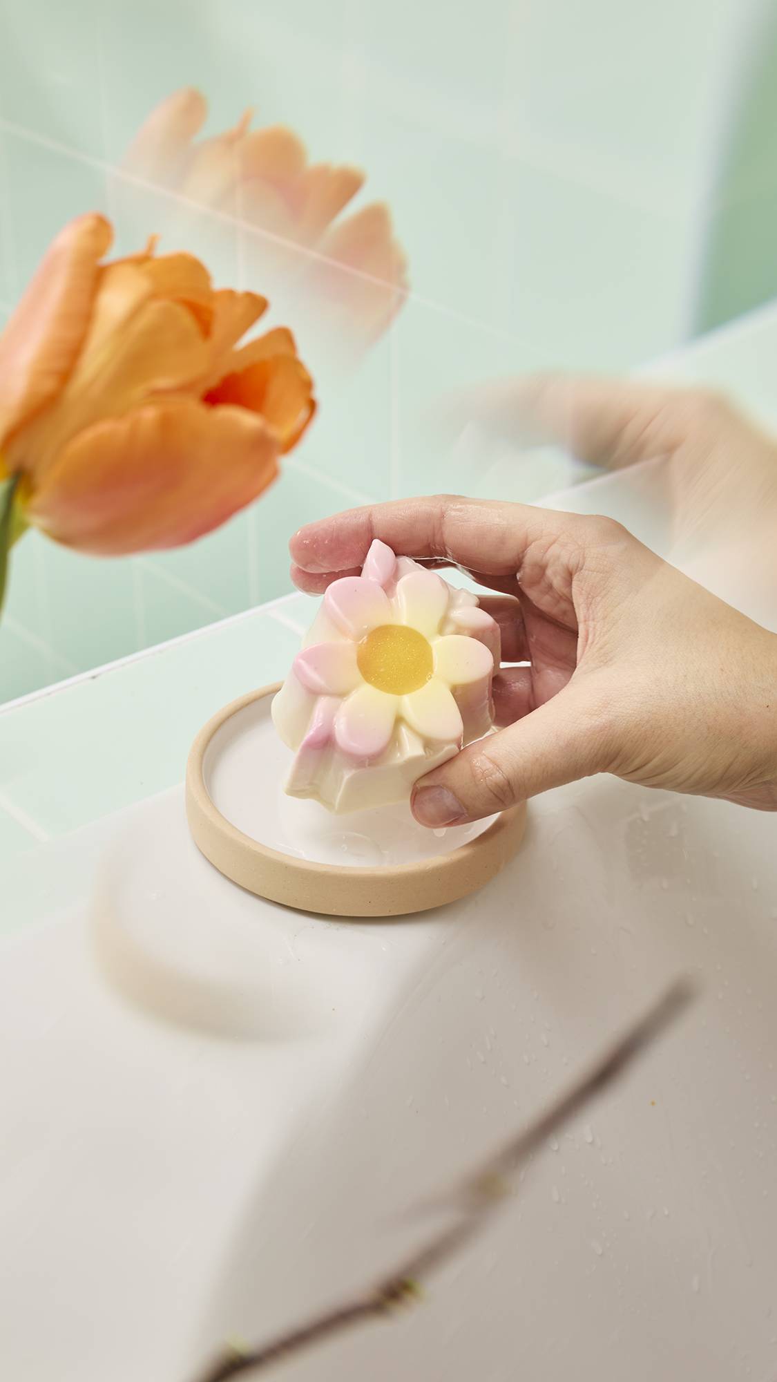 A hand places the flower-shaped "Making Daisy Chains" bar onto a small beige dish on a white ledge. The product has white petals with pink tips and a yellow center, framed by a blurred orange tulip and green tiles.