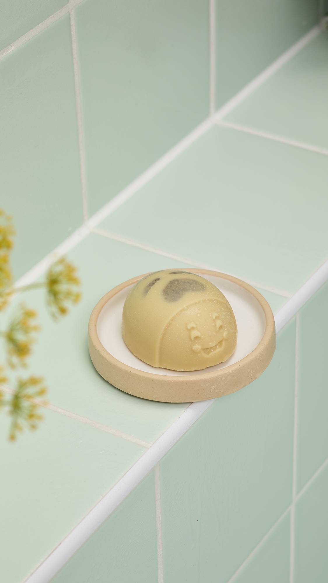 A cream "Mama-chino Scrubee" body butter bar sits on a beige dish against mint green tiles. The bee-shaped bar features a smiling face and dark stripes, framed by blurred yellow flowers.