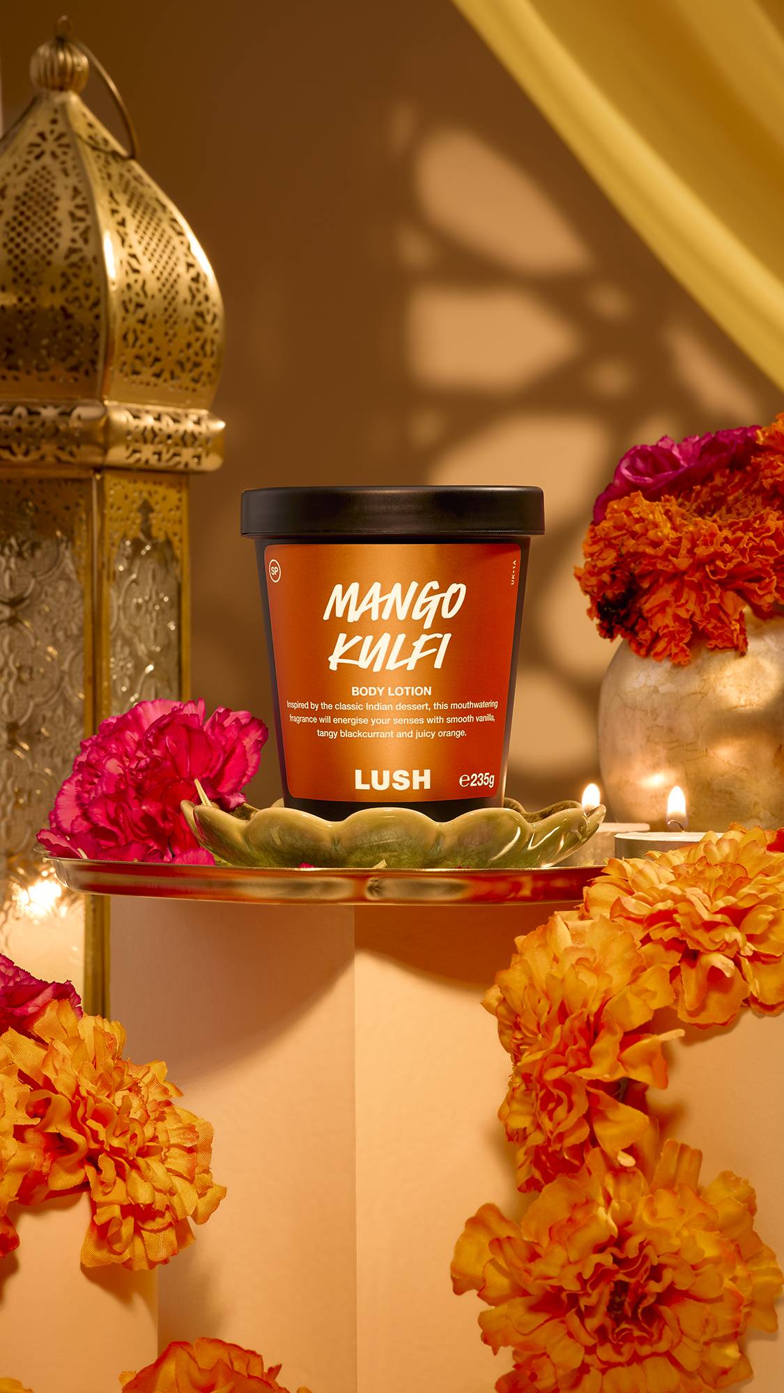 A black pot of Lush Mango Kulfi body lotion sits on a golden plate surrounded by bright marigold flowers, magenta carnations, lit tealights, and an ornate brass lantern in a warm Diwali setting.
