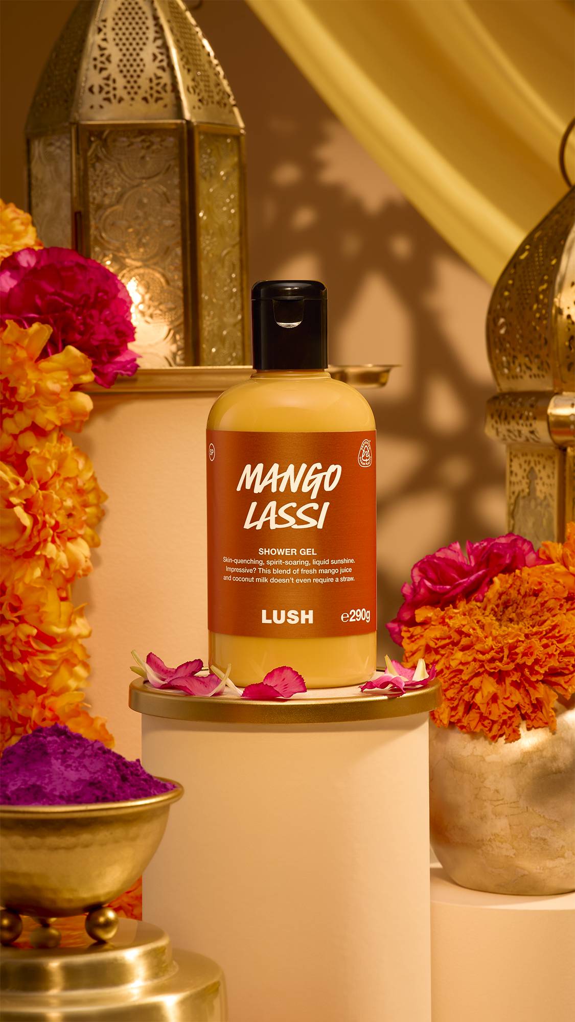 A bottle of Lush Mango Lassi shower gel displayed on a small pedestal surrounded by pink petals, vibrant marigold flowers, brass lanterns, and a bowl of purple powder pigment in a warm Diwali setup.