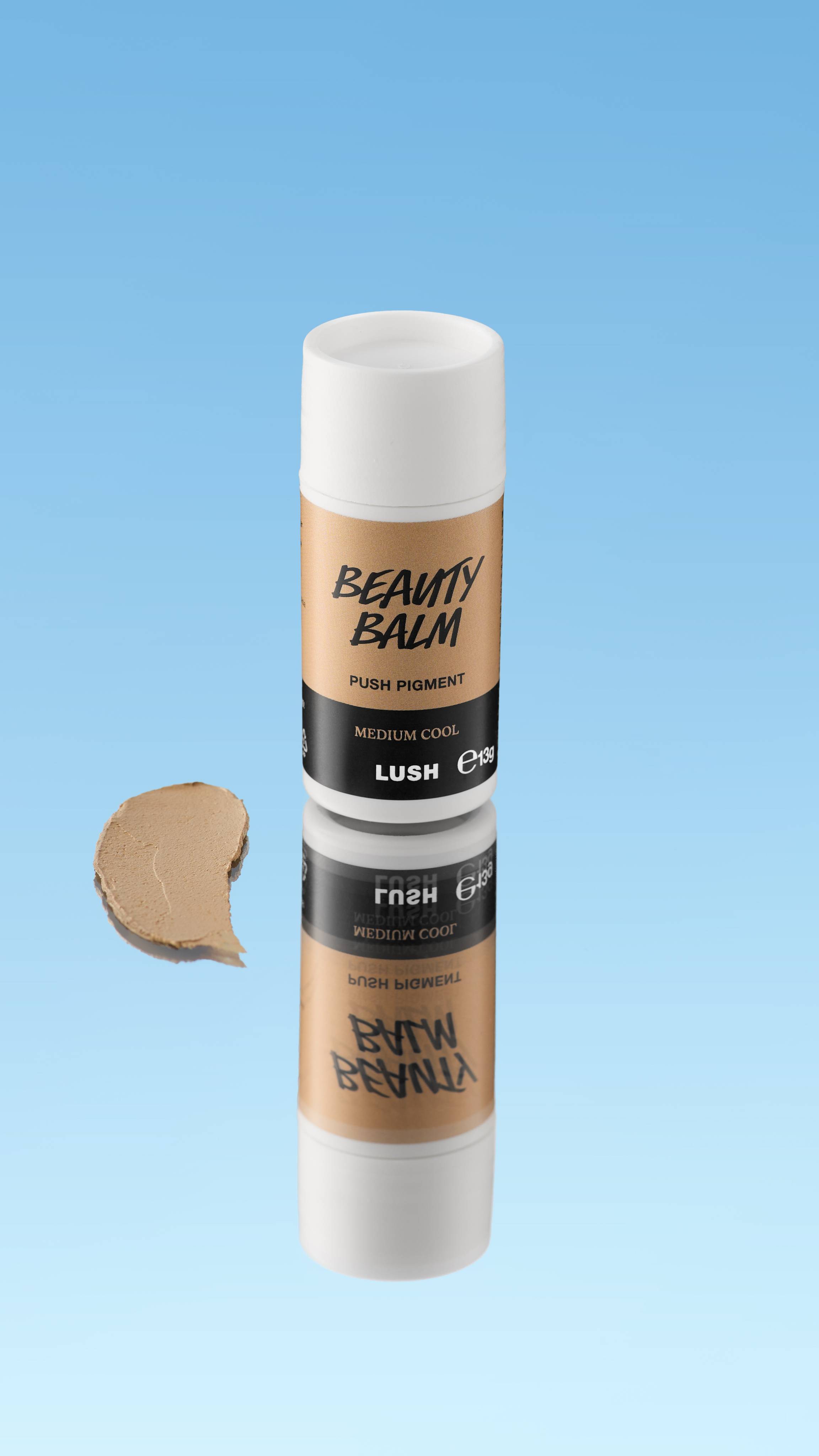 An angled product shot of a white and taupe-brown LUSH Beauty Balm Push Pigment stick in the shade Medium Cool. A thick swatch of the cool-toned medium brown product rests next to the reflected stick, all against a bright blue background.