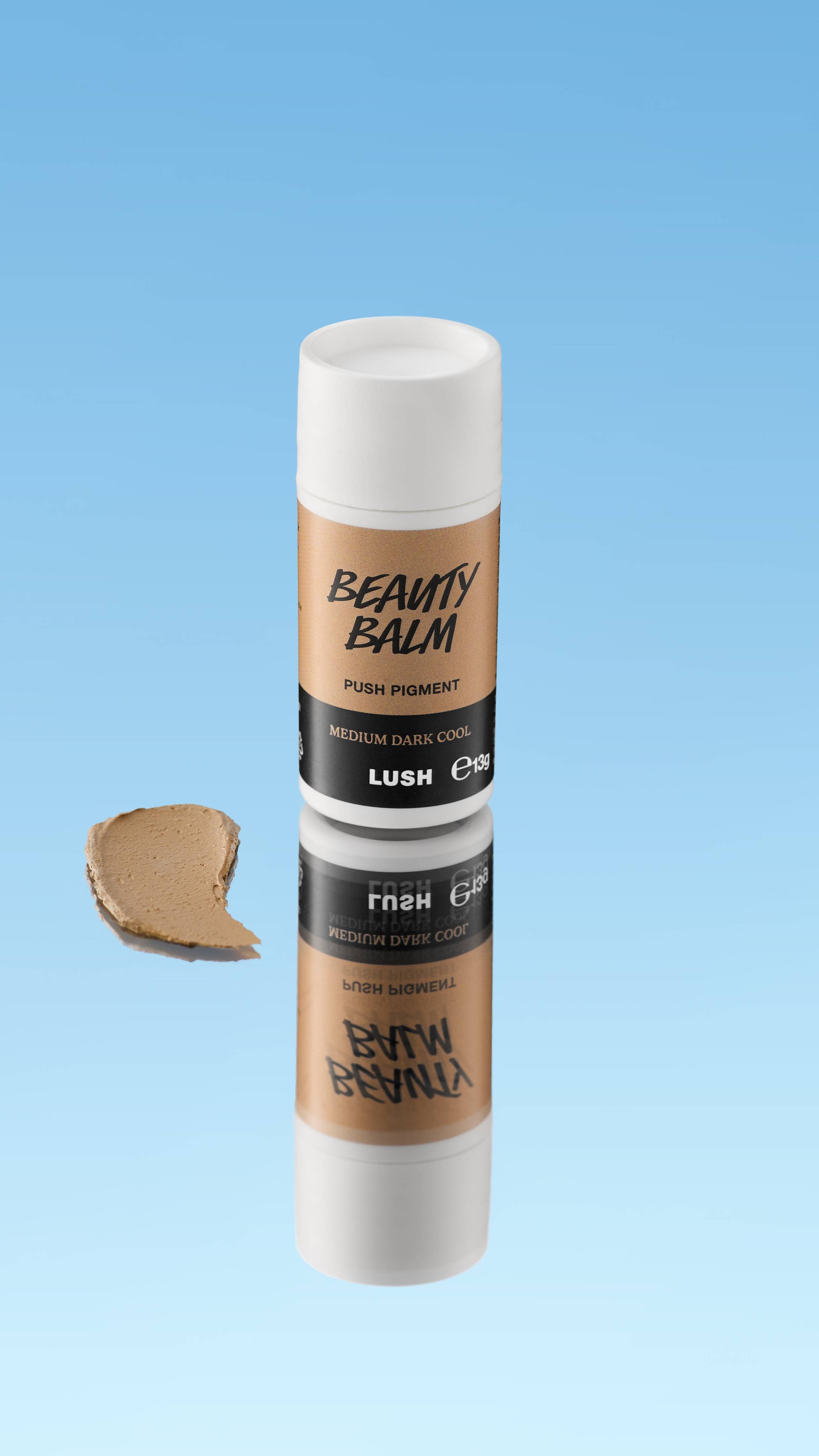 An angled product shot of a white and olive-brown LUSH Beauty Balm Push Pigment stick in the shade Medium Dark Cool. A thick swatch of the cool-toned brown product rests next to the reflected stick, all against a bright blue background.