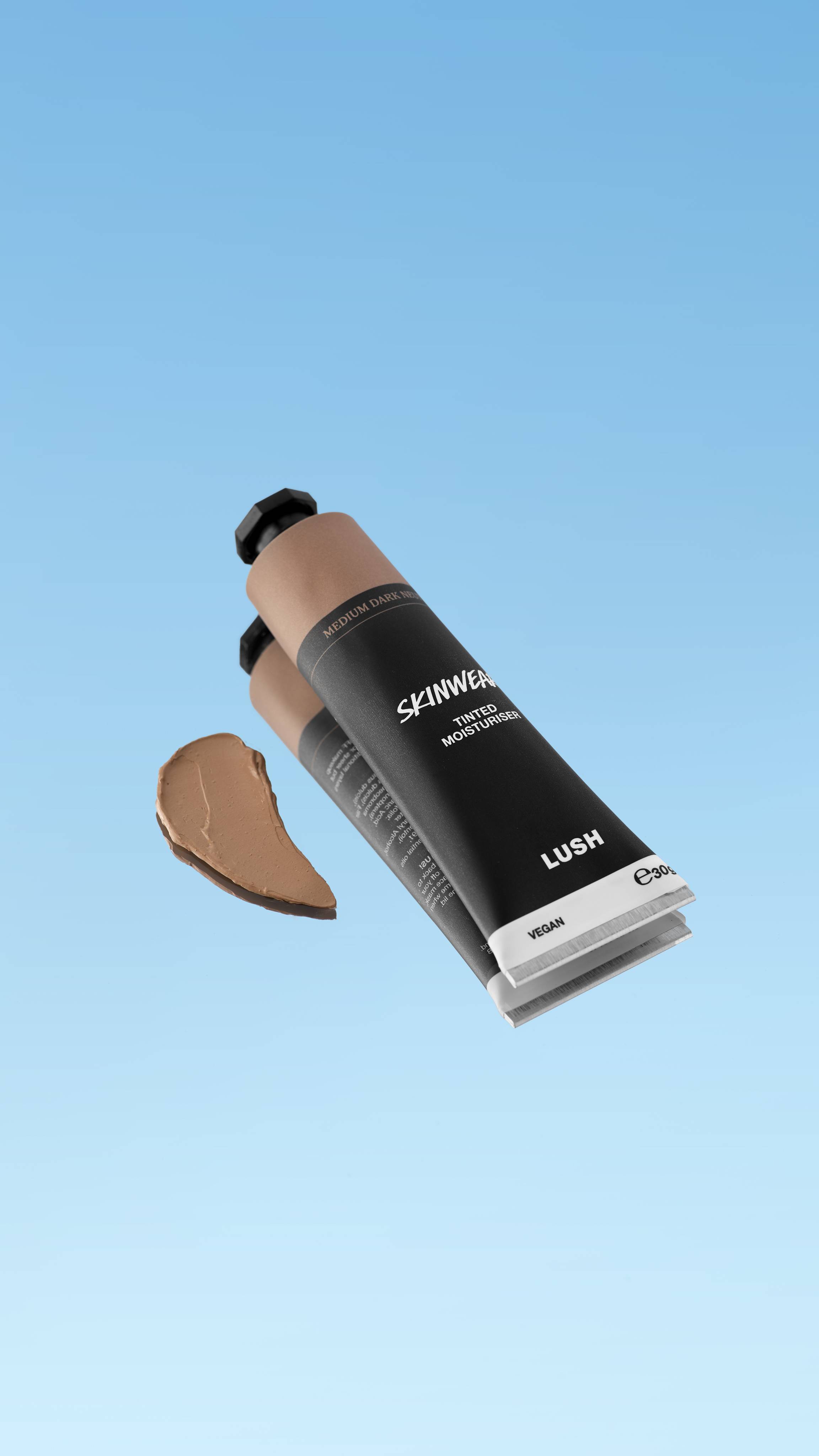 A product shot showing the black tube of Skinwear Tinted Moisturiser in the Medium Dark Neutral shade, floating against a blue gradient background. A dollop of the warm, medium beige-tan cream is swatched to the left of the tube.
