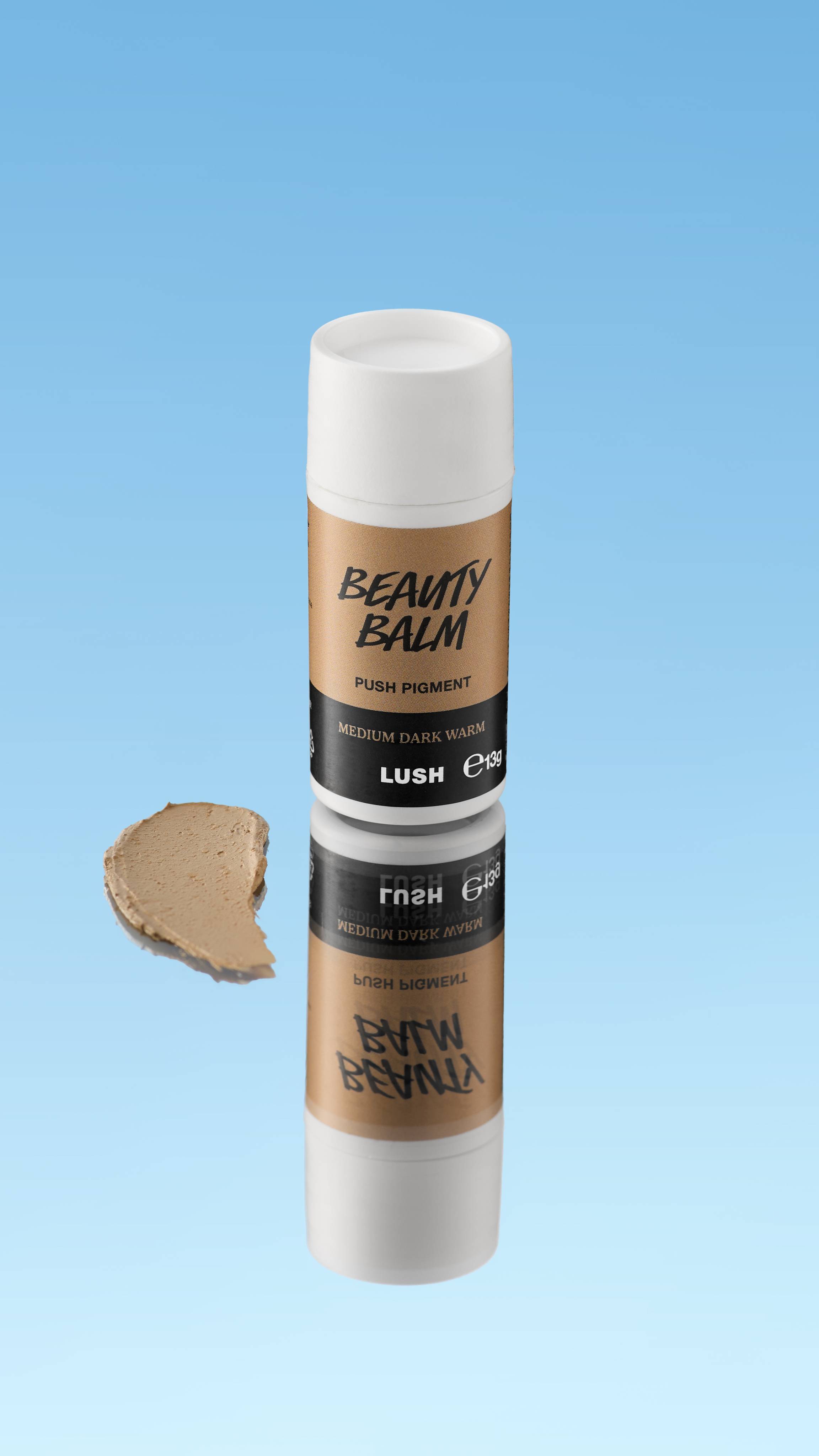 An angled product shot of a white and golden-brown LUSH Beauty Balm Push Pigment stick in the shade Medium Dark Warm. A thick swatch of the warm brown product rests next to the reflected stick, all against a bright blue background.