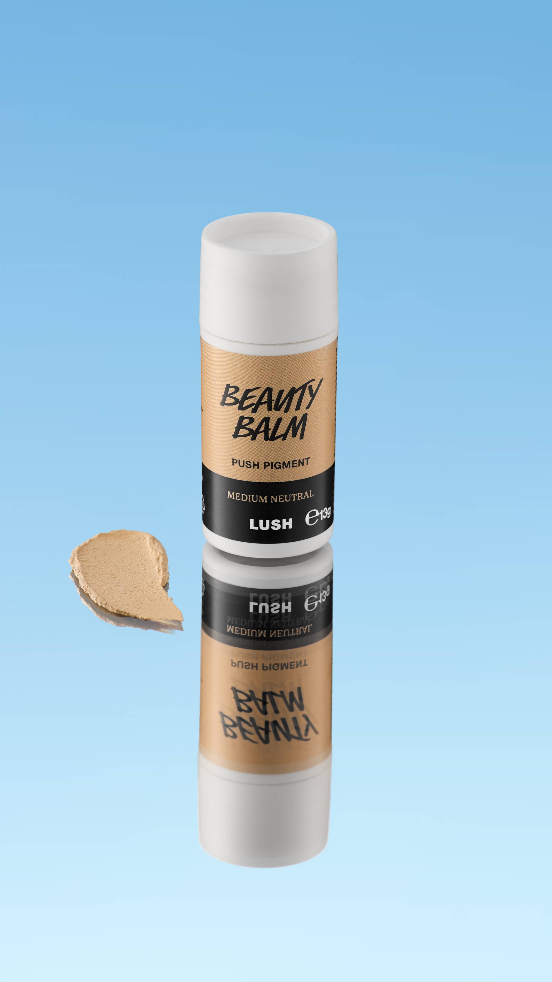 An angled product shot of a white and tan LUSH Beauty Balm Push Pigment stick in the shade Medium Neutral. A thick swatch of the neutral medium beige product rests next to the reflected stick, all against a bright blue background.