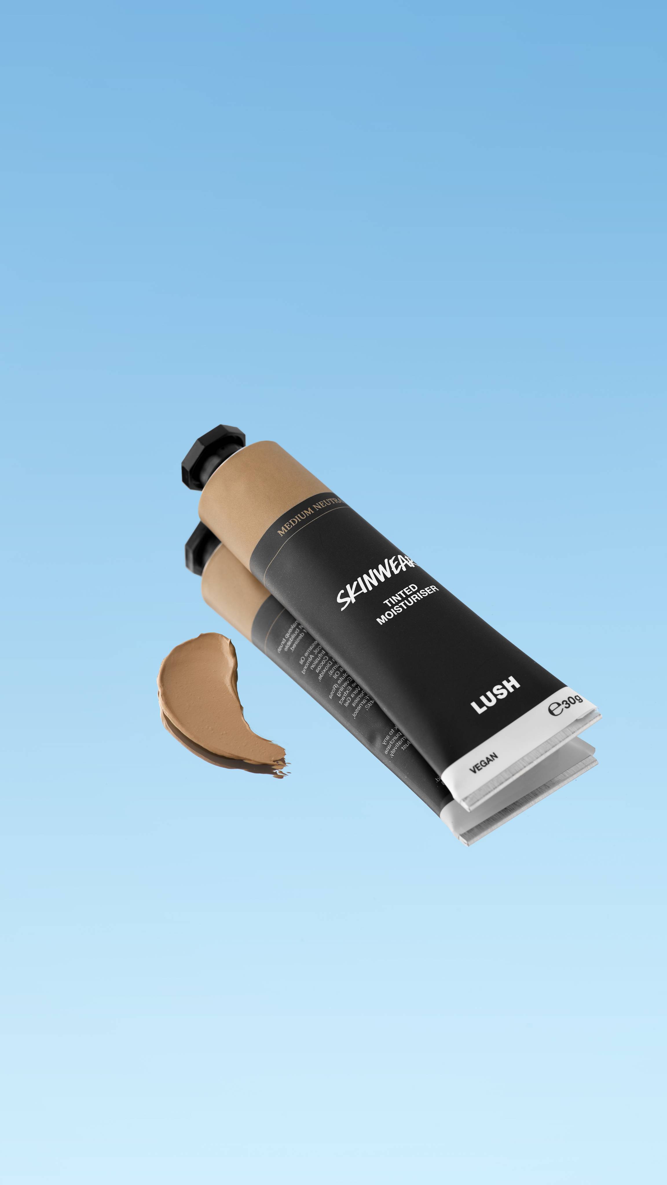 A product shot showing the black tube of Skinwear Tinted Moisturiser in the Medium Neutral shade, floating against a blue gradient background. A dollop of the light-to-medium golden beige cream is swatched to the left of the tube.