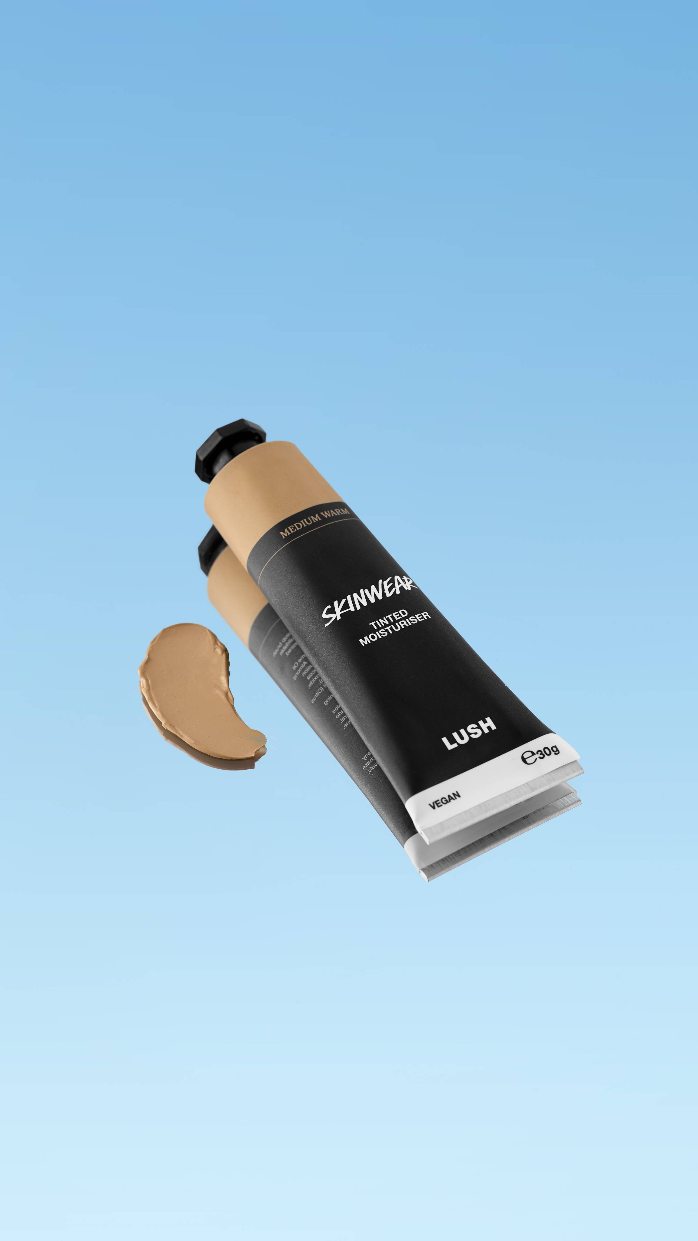 A product shot showing the black tube of Skinwear Tinted Moisturiser in the Medium Warm shade, floating against a blue gradient background. A dollop of the medium golden-yellow beige cream is swatched to the left of the tube.